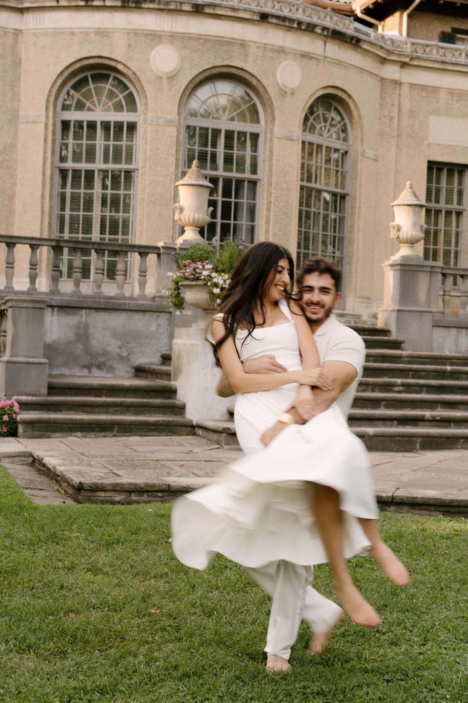 Man lifting and spinning laughing woman in white dress in front of elegant stone mansion exterior.