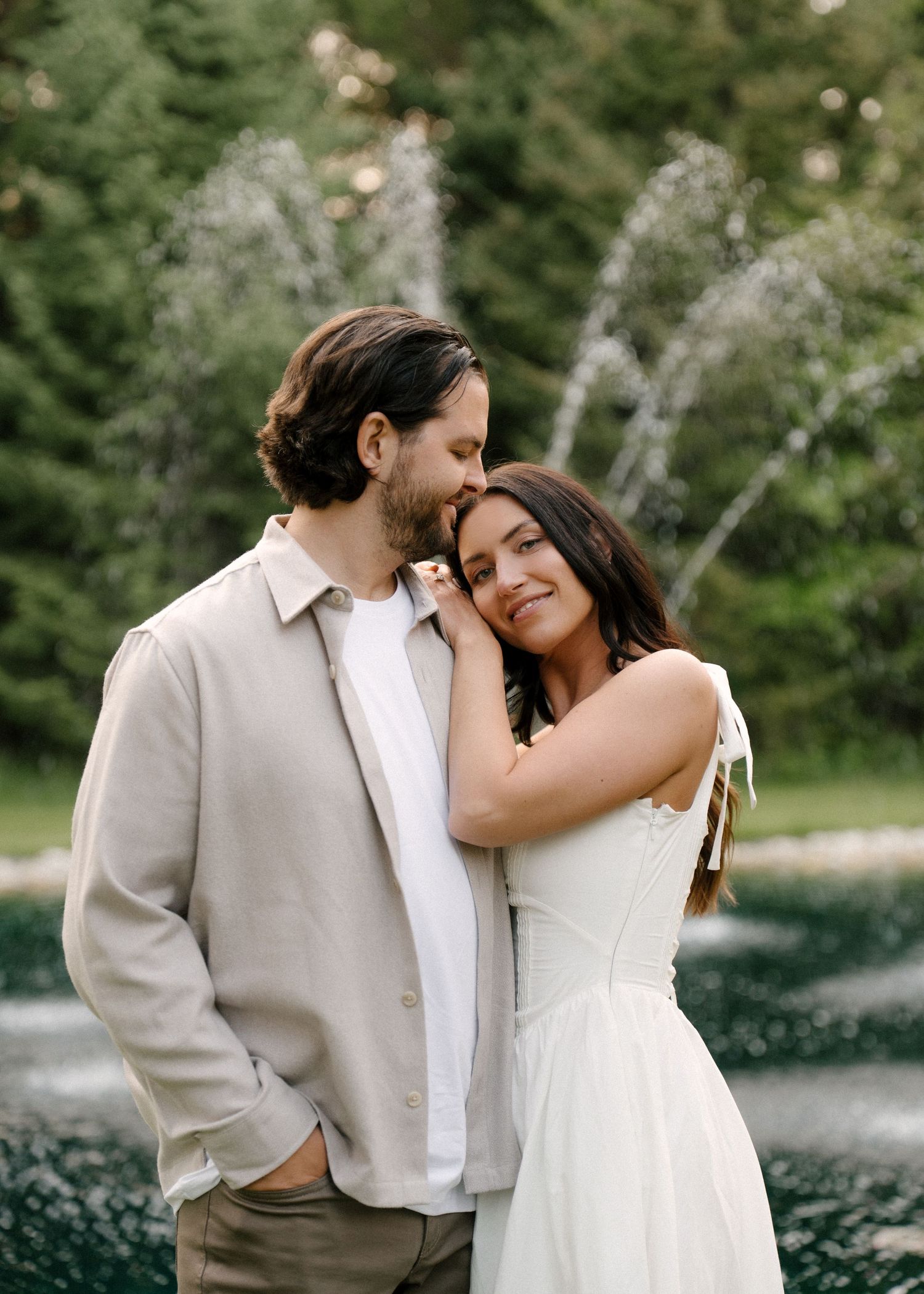 Couple embracing near a fountain in a lush garden, man in beige jacket nuzzling woman in white dress.