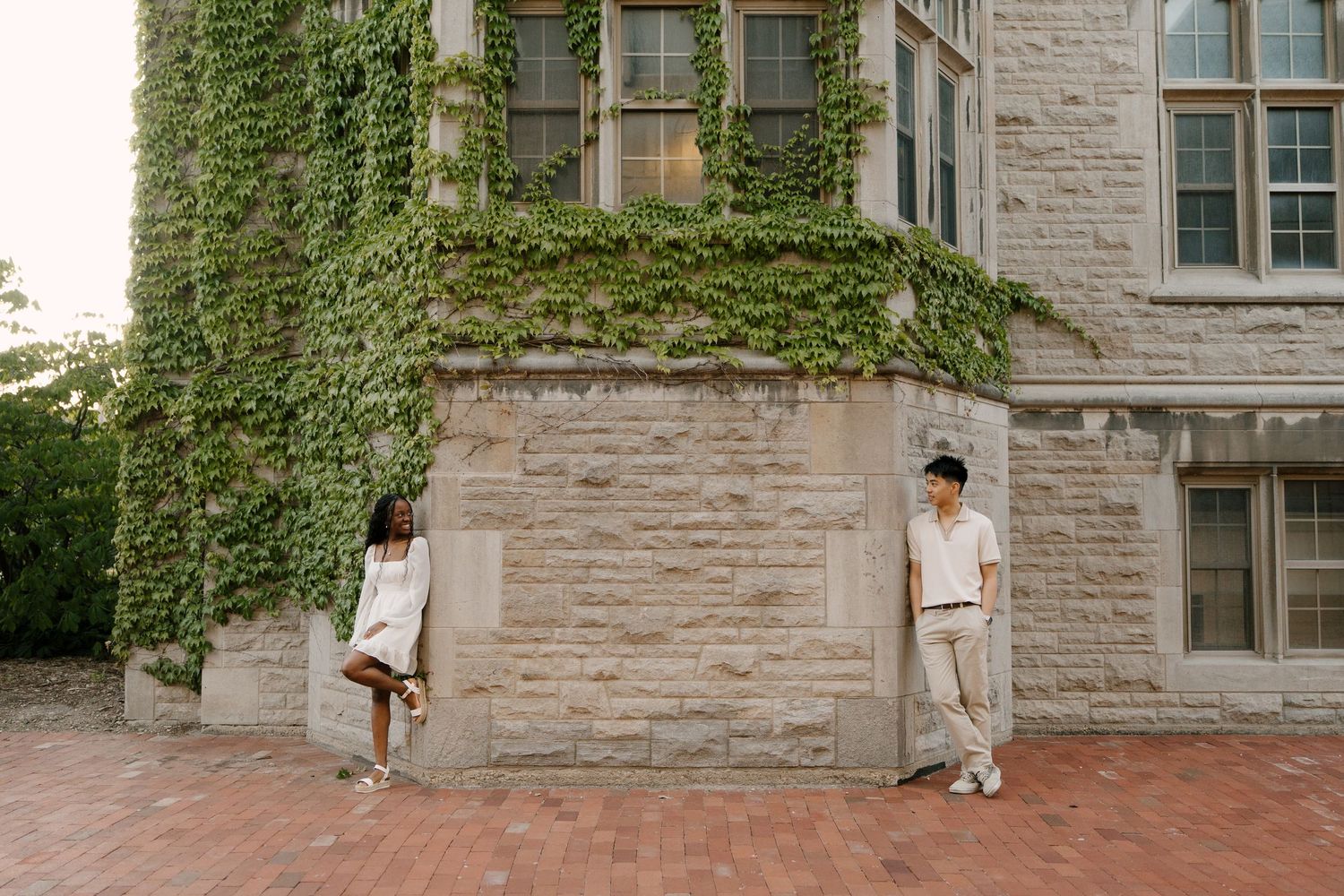 Interracial couple leaning against an ivy-covered stone building corner, posing apart on a brick plaza.