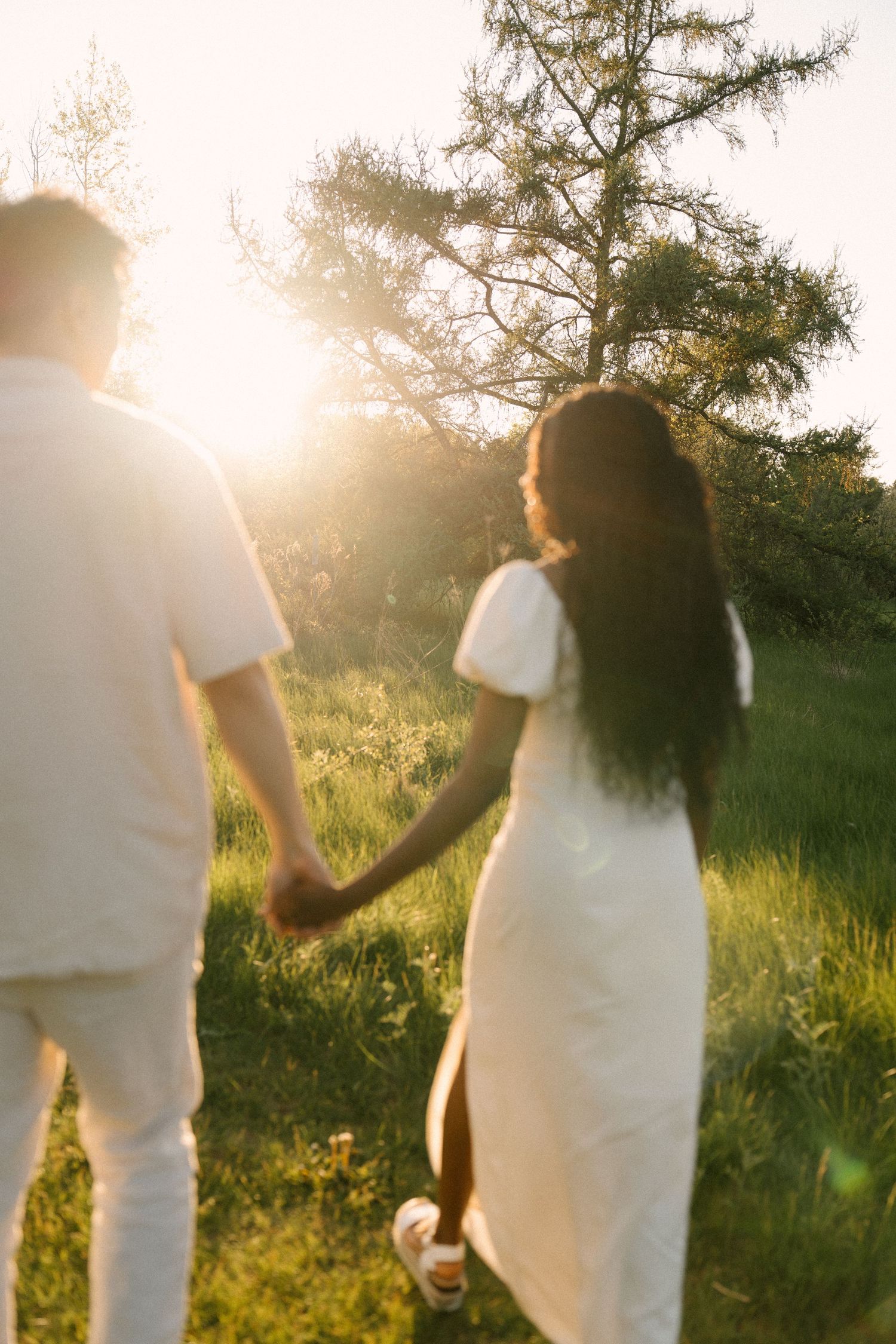 Couple holding hands walking toward glowing sunset in lush green meadow.
