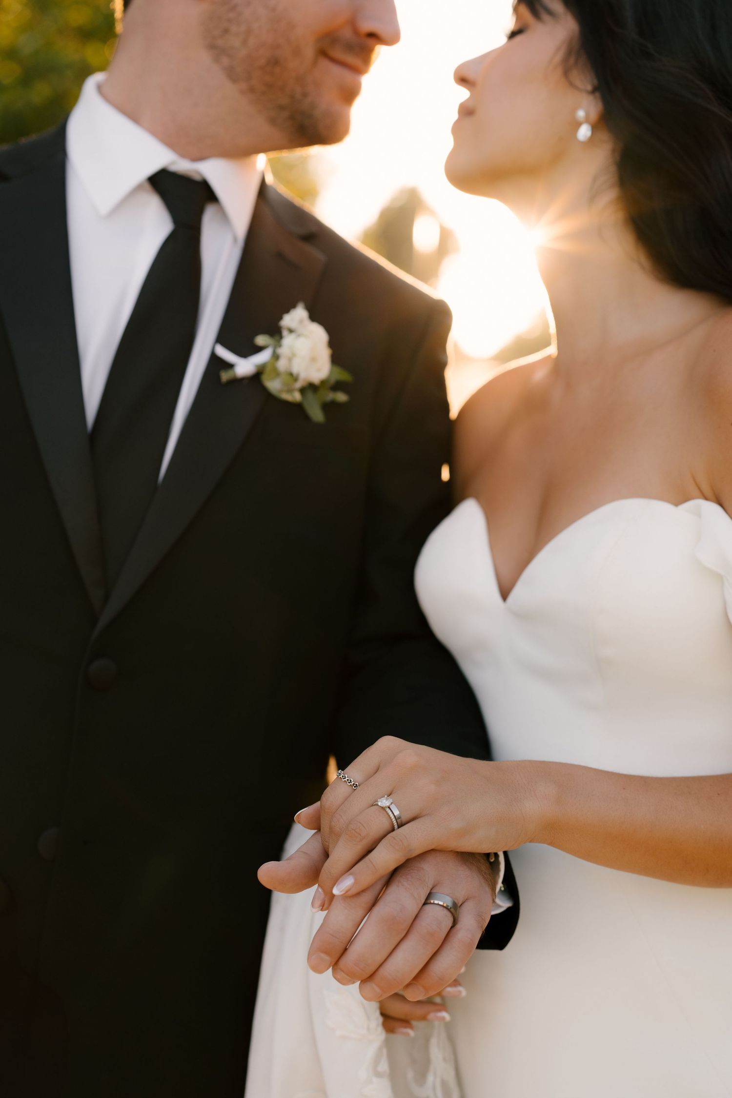 Close-up of bride and groom holding hands showing wedding rings, groom wearing white boutonniere in warm sunlight.