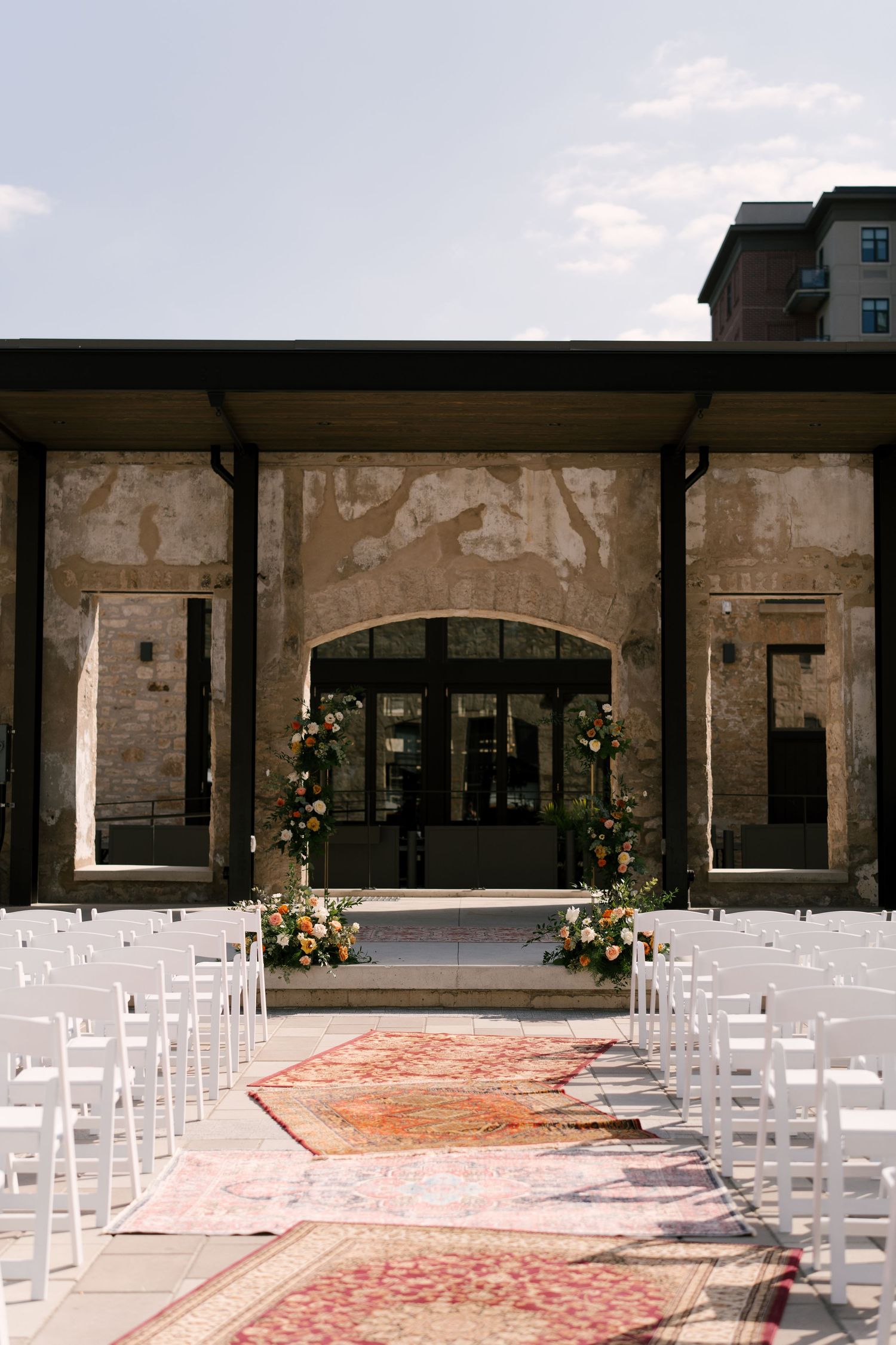 Elegant outdoor Tapestry Hall wedding ceremony setup with white chairs, colourful rugs as aisle runner, and floral arch at altar.