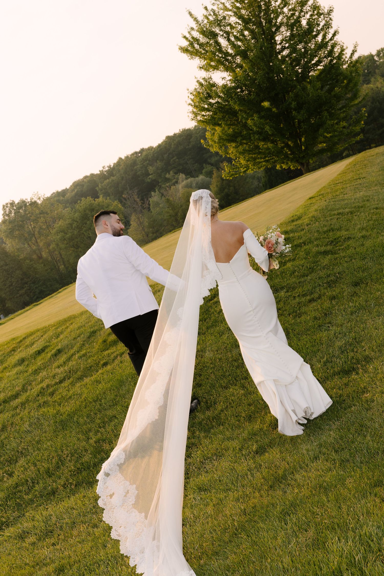 Bride and groom walking up grassy hill at sunset, cathedral veil trailing behind white fitted wedding gown at Whistle Bear venue.
