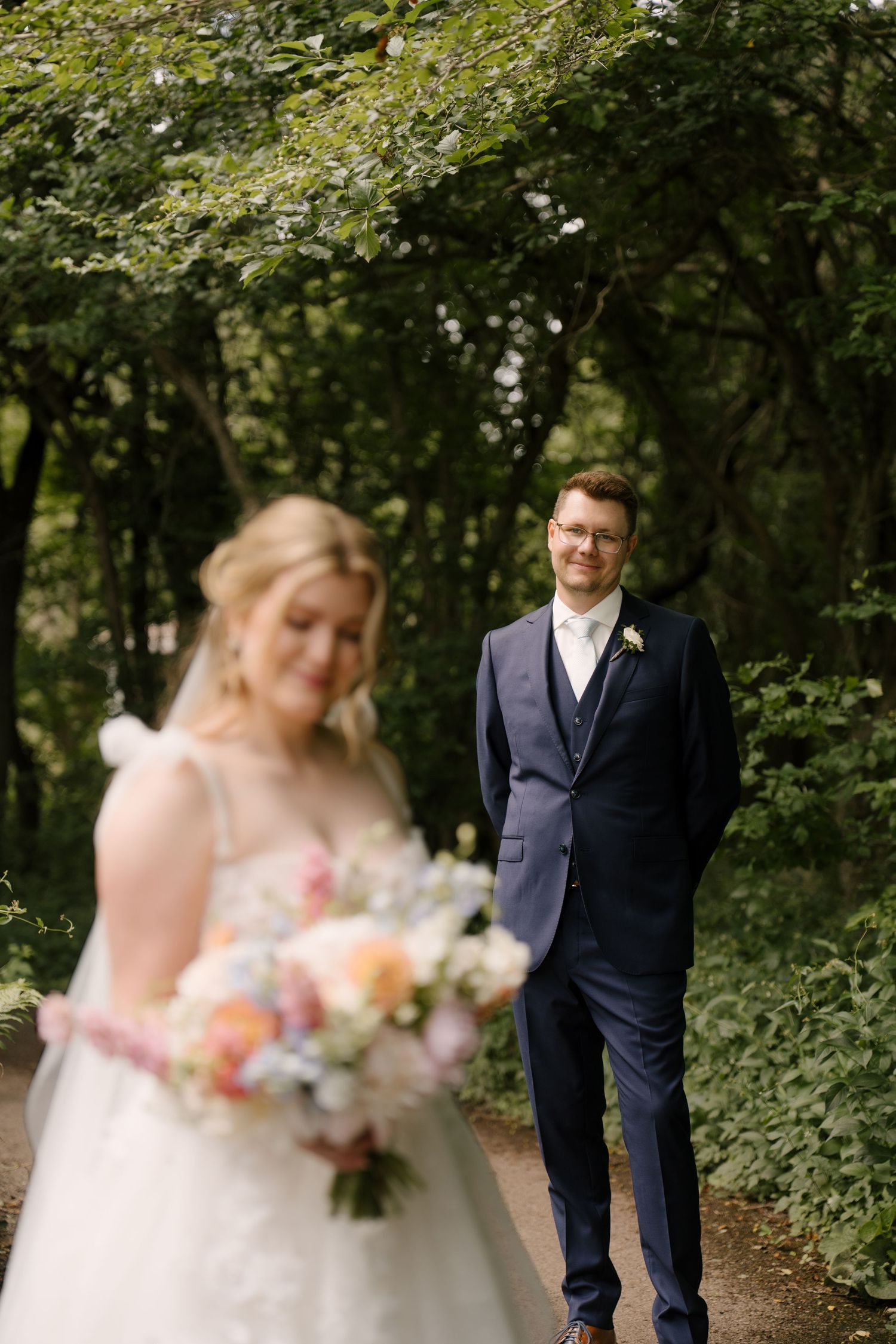 Groom in navy suit smiles admiringly at bride holding colourful bouquet in lush garden at St Marys Golf Club.