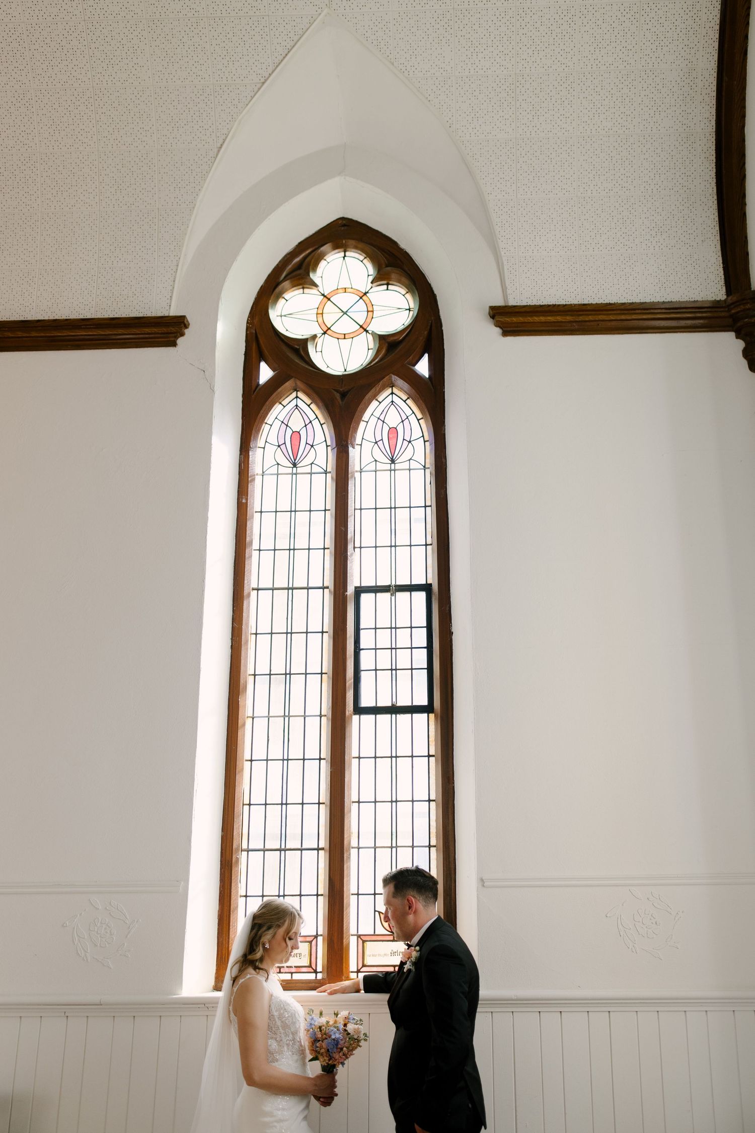 Bride and groom share an intimate moment before a stunning arched stained glass church window.