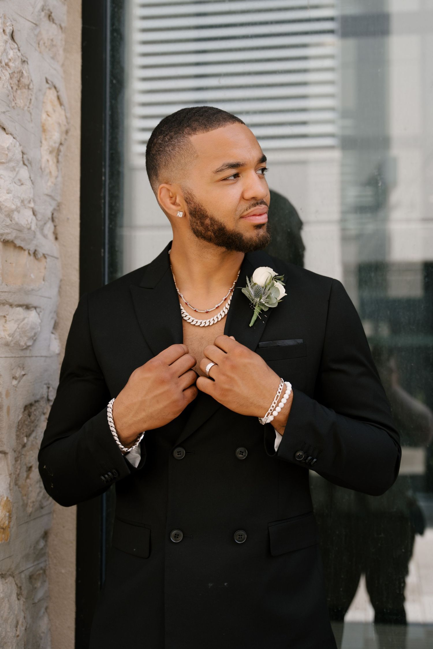 Dapper groom in a black suit adjusts his lapel while wearing a silver chain and white boutonniere.
