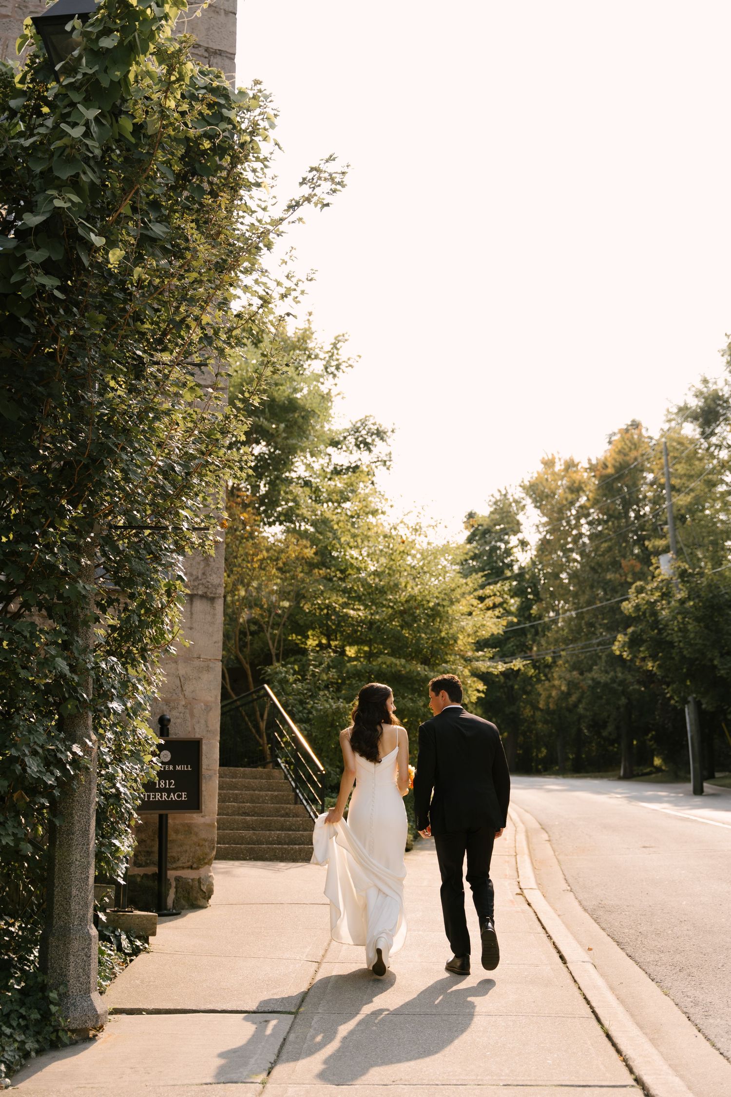 Bride and groom walking hand in hand down a sunlit sidewalk surrounded by lush green trees.