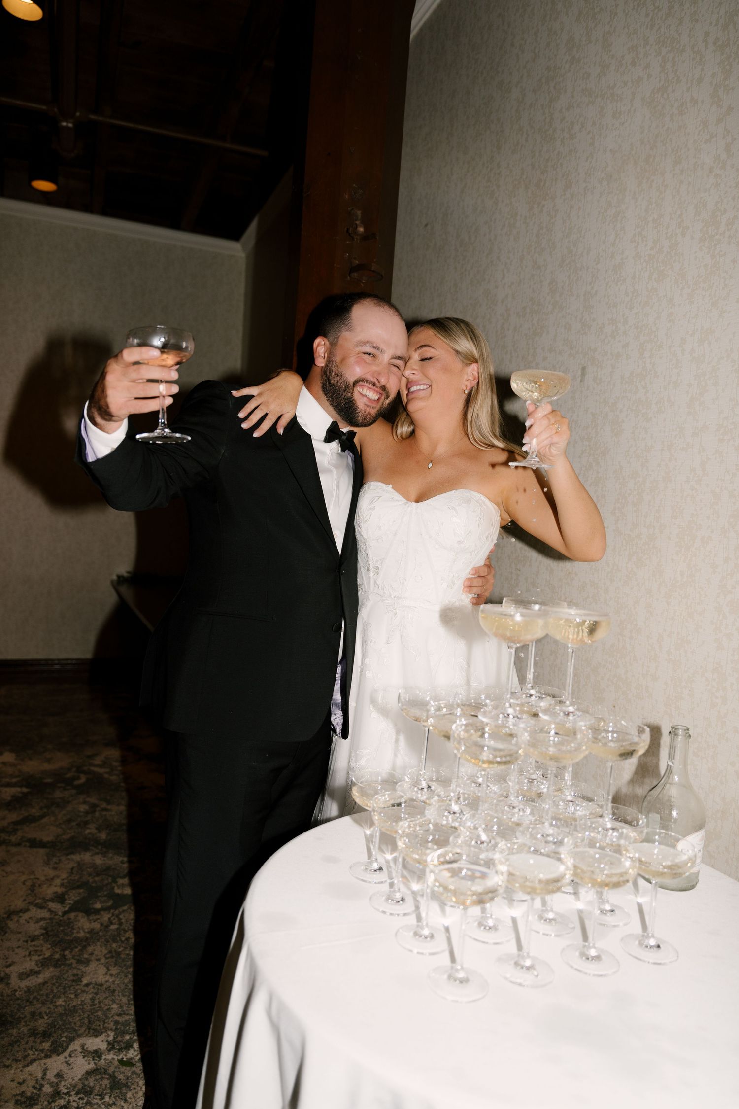 Smiling newlyweds toast with champagne coupes beside a sparkling champagne tower at their Ancaster Mill wedding reception.