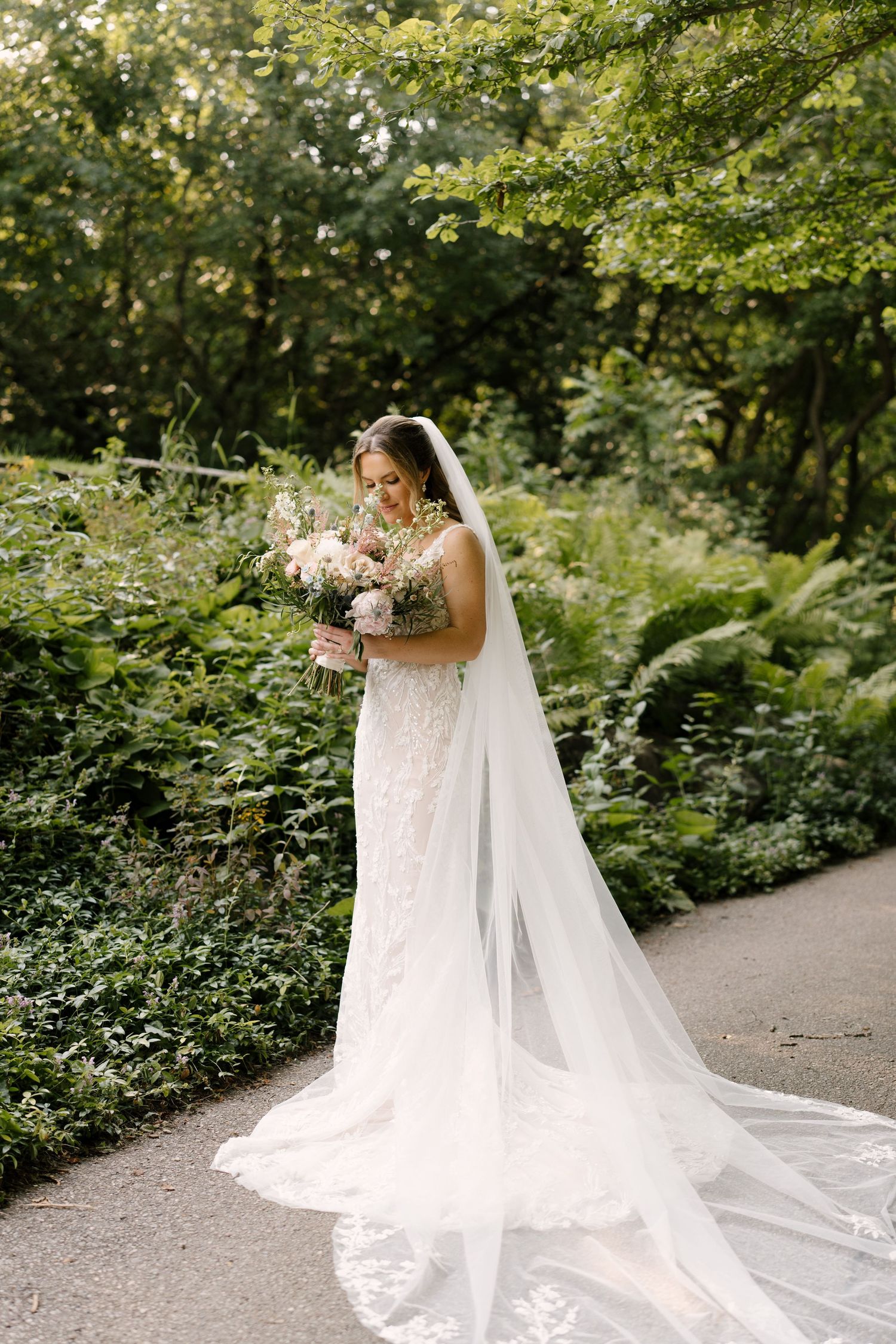 Bride in lace gown and long veil admiring her blush bouquet on a garden path surrounded by greenery at St Marys venue.