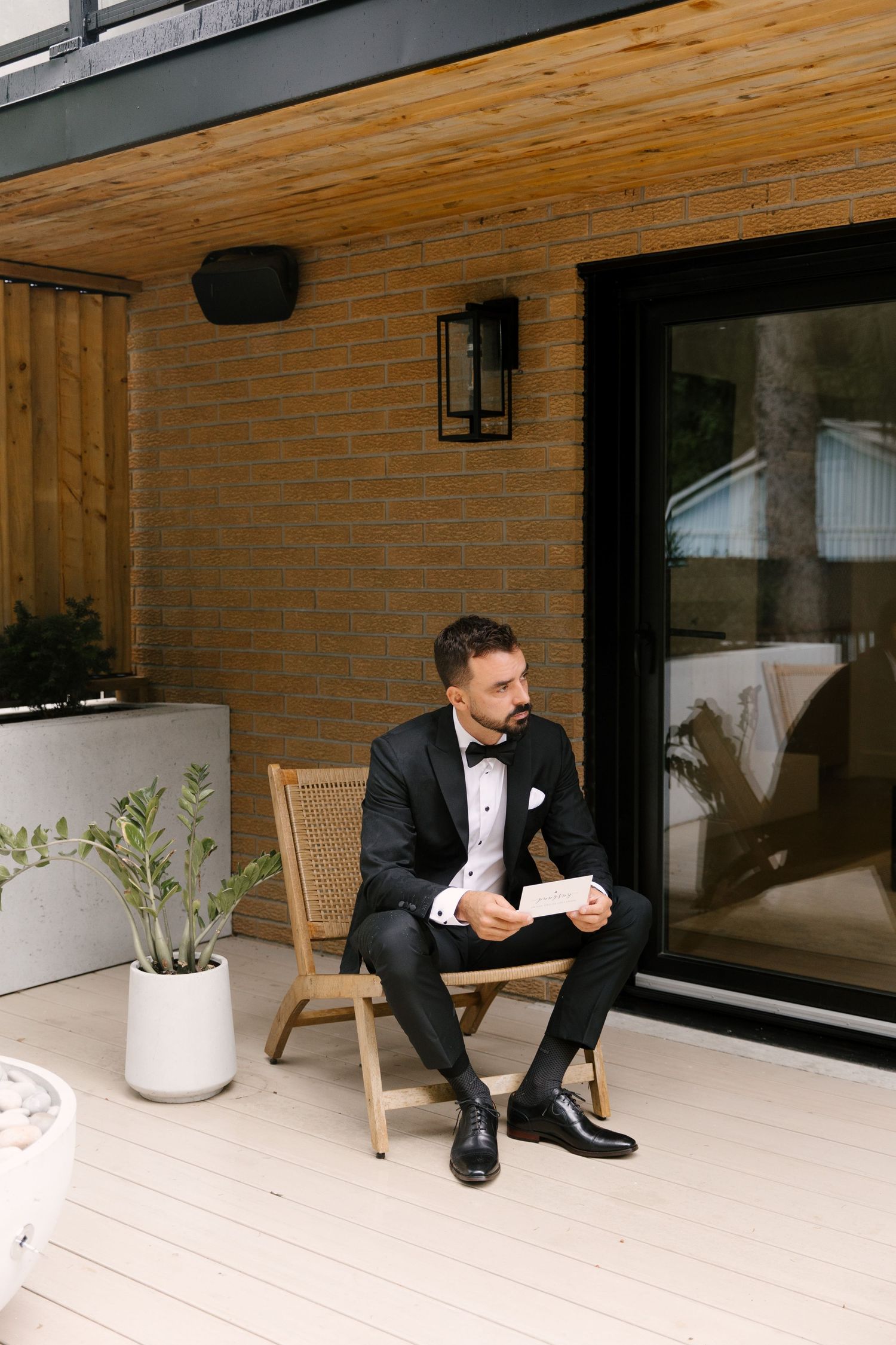 Dapper groom in black tuxedo sits on patio chair holding a card outside brick home.