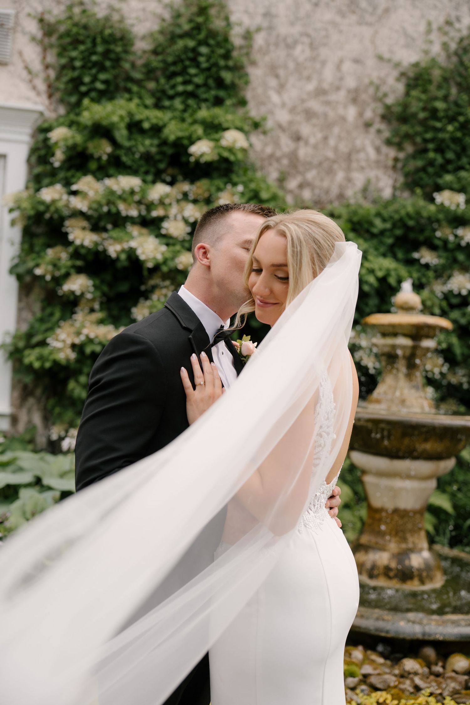 Groom tenderly kisses bride near stone fountain as her veil flows in the breeze at Inn On The Twenty.