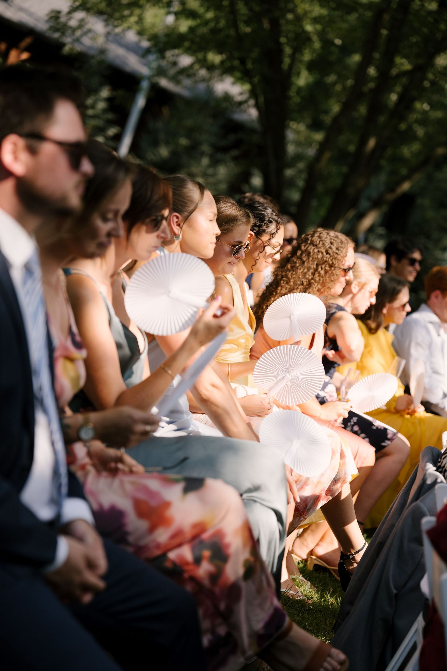 Wedding guests seated outdoors fanning themselves with white paper fans during a sunny outdoor ceremony.