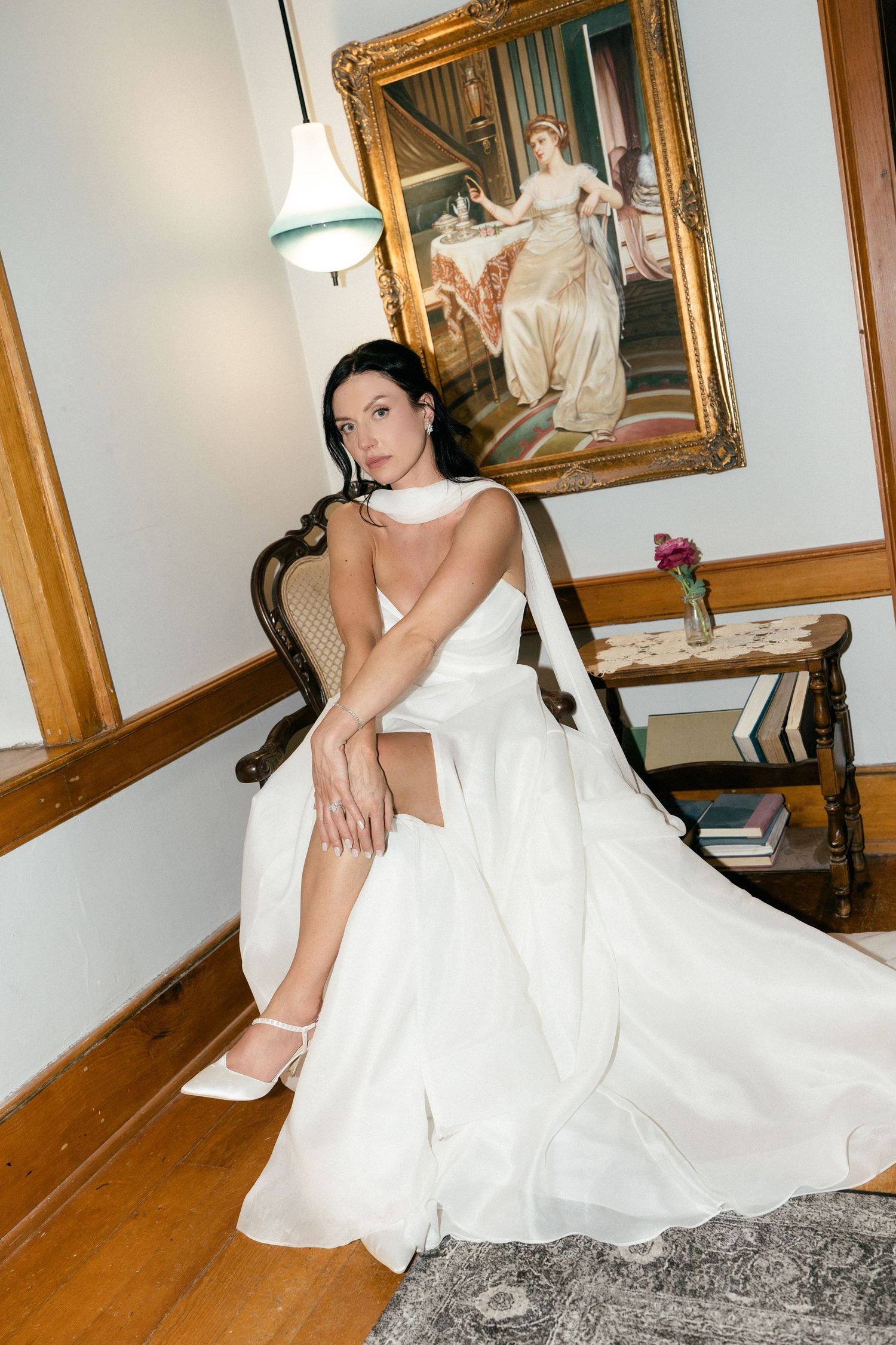 Bride in white strapless ball gown sitting elegantly in vintage chair beside classic painting.