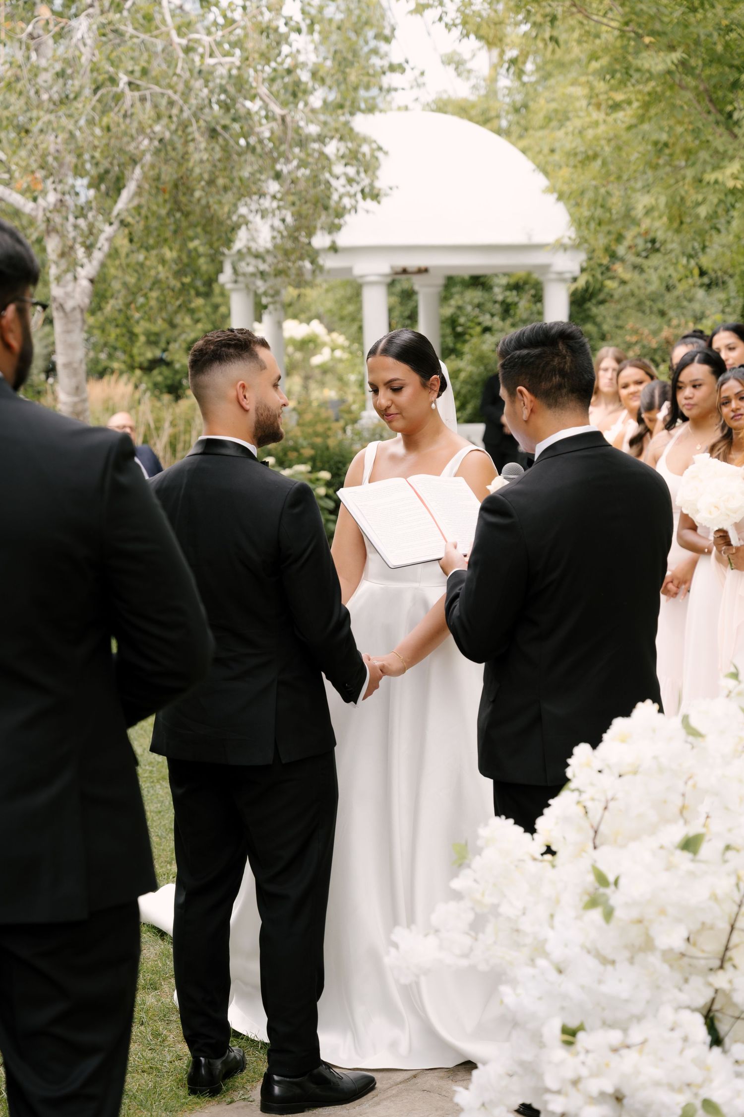 Bride and groom exchange vows at outdoor garden ceremony surrounded by white floral arrangements and wedding party.