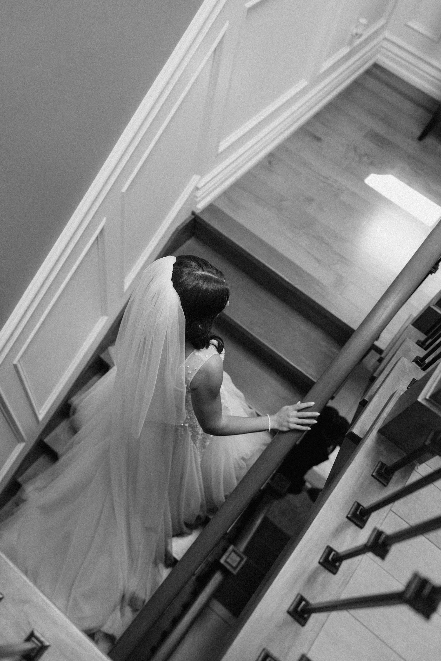 Black and white photo of a bride in a white gown and veil descending an elegant staircase on her wedding day.