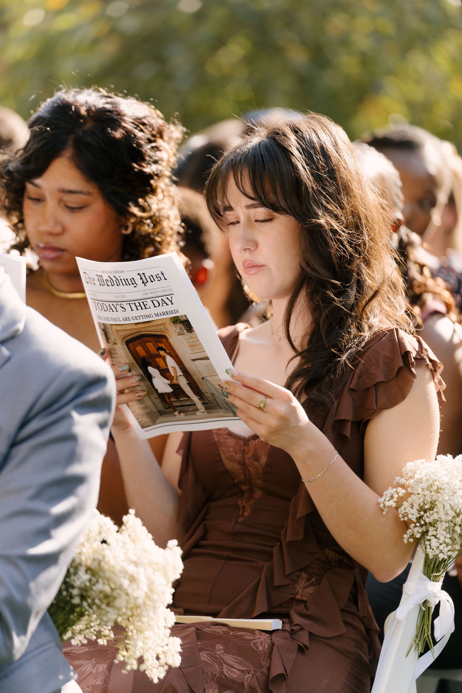 Emotional wedding guest reading a newspaper-style wedding program during an outdoor ceremony.