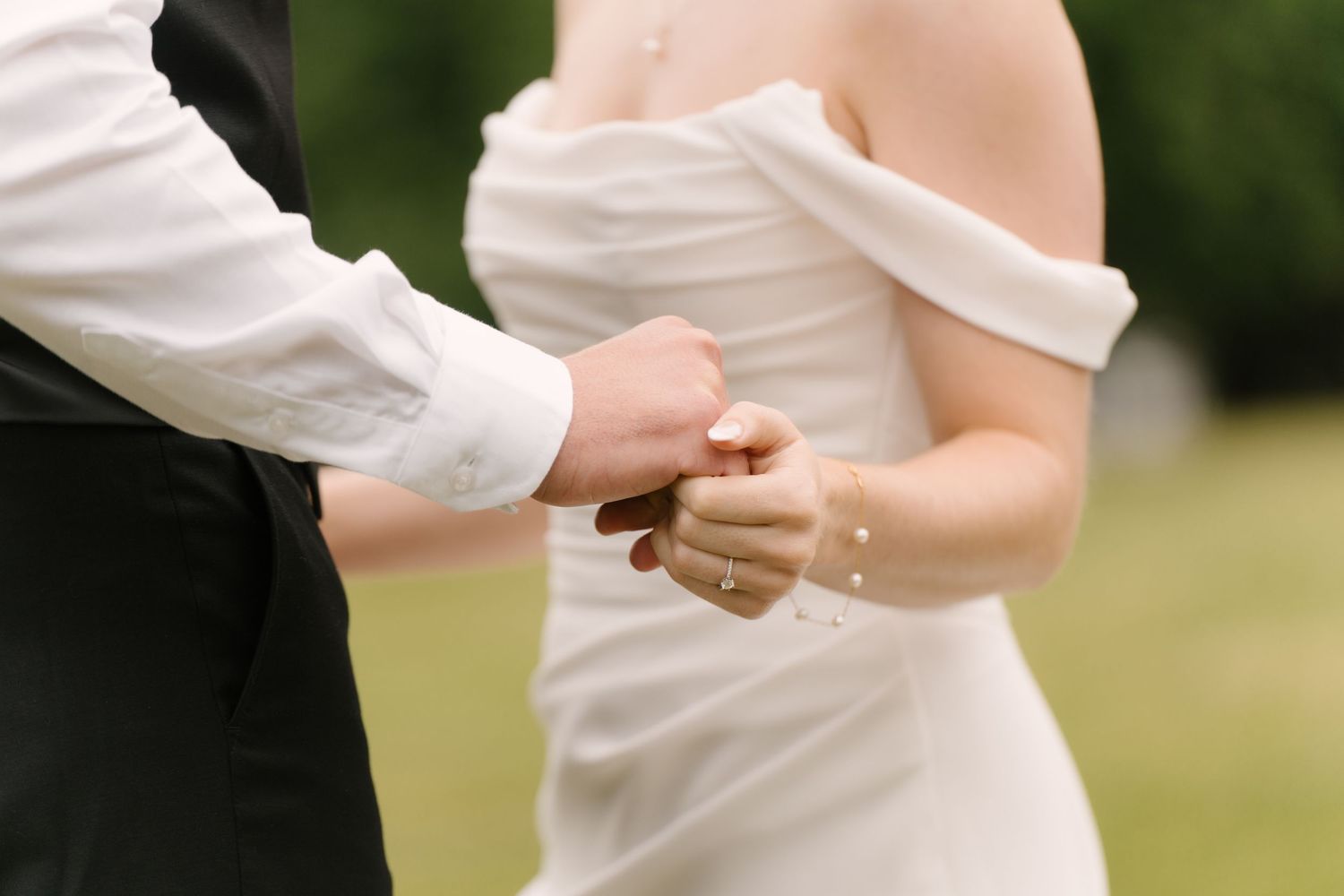 Groom holding bride's hand outdoors, showcasing her elegant off-shoulder white gown.