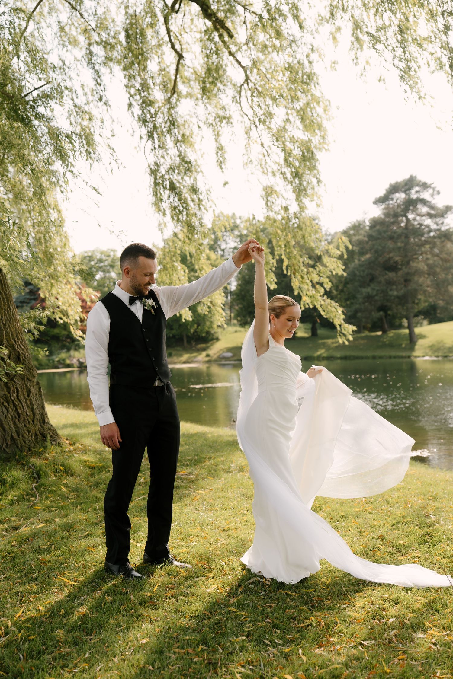 Groom twirls bride in a flowing white gown under a willow tree by a peaceful lake.