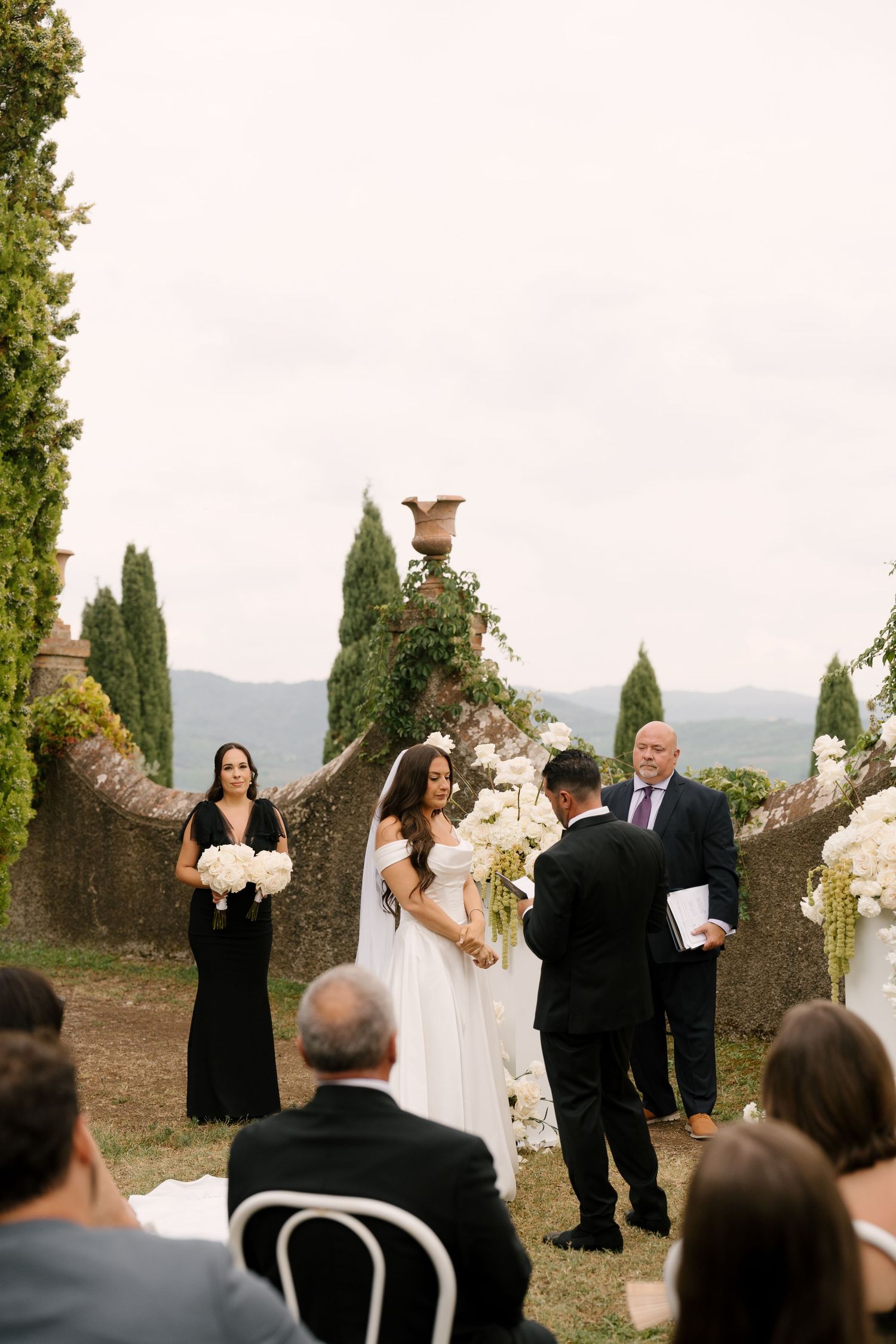 Couple holds hands during outdoor Tuscan wedding ceremony at Italian villa surrounded by cypress trees and white flowers.