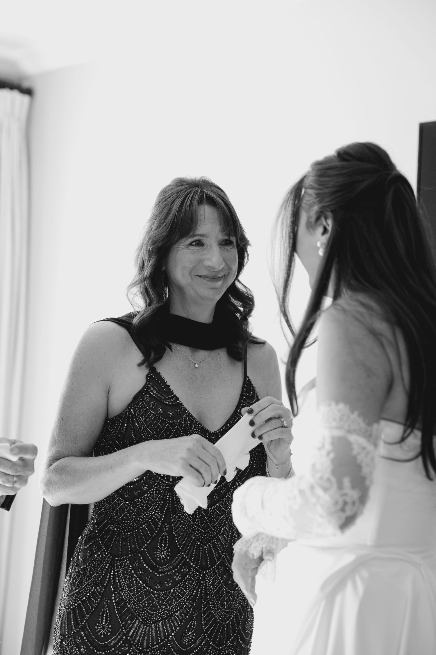 Emotional woman in beaded black dress smiles at bride during wedding preparations, black and white.