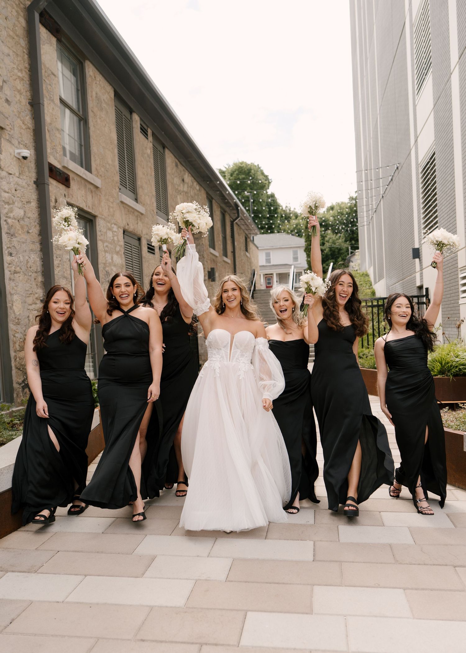 Bride in white gown celebrates with six bridesmaids in black dresses raising bouquets outdoors at Tapestry Hall wedding venue.