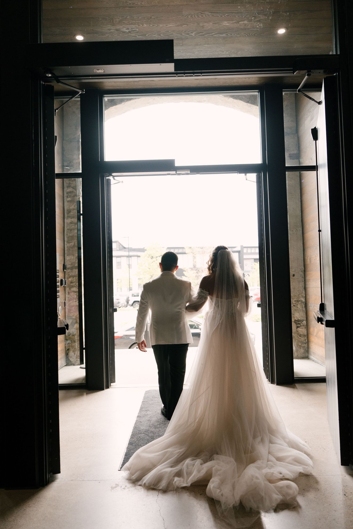 Bride and groom silhouetted holding hands while walking through grand open doors toward bright natural light outside at Tapestry Hall.