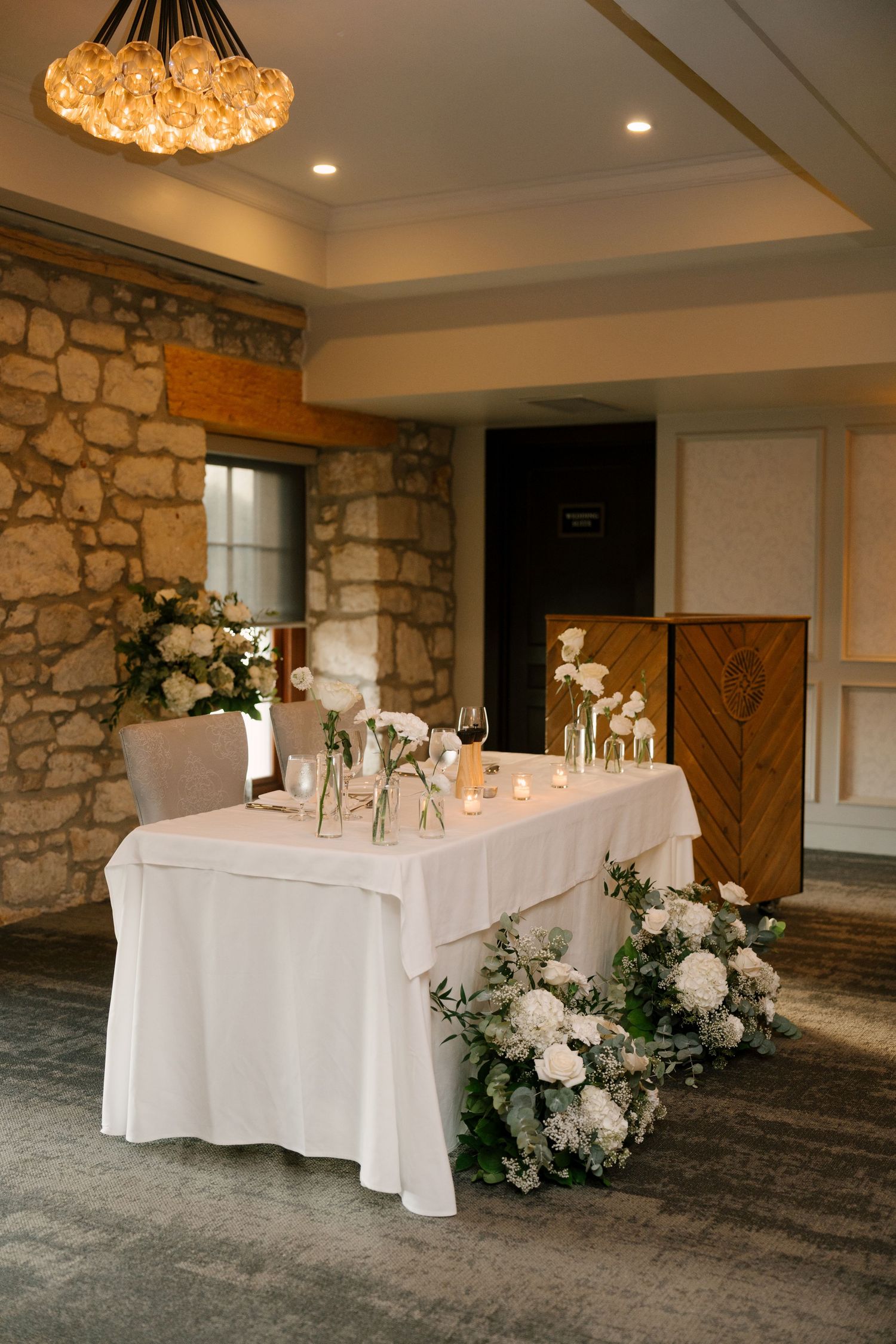 Romantic sweetheart table with white florals, greenery, candles, and stone wall backdrop at Cambridge Mill.wedding reception.