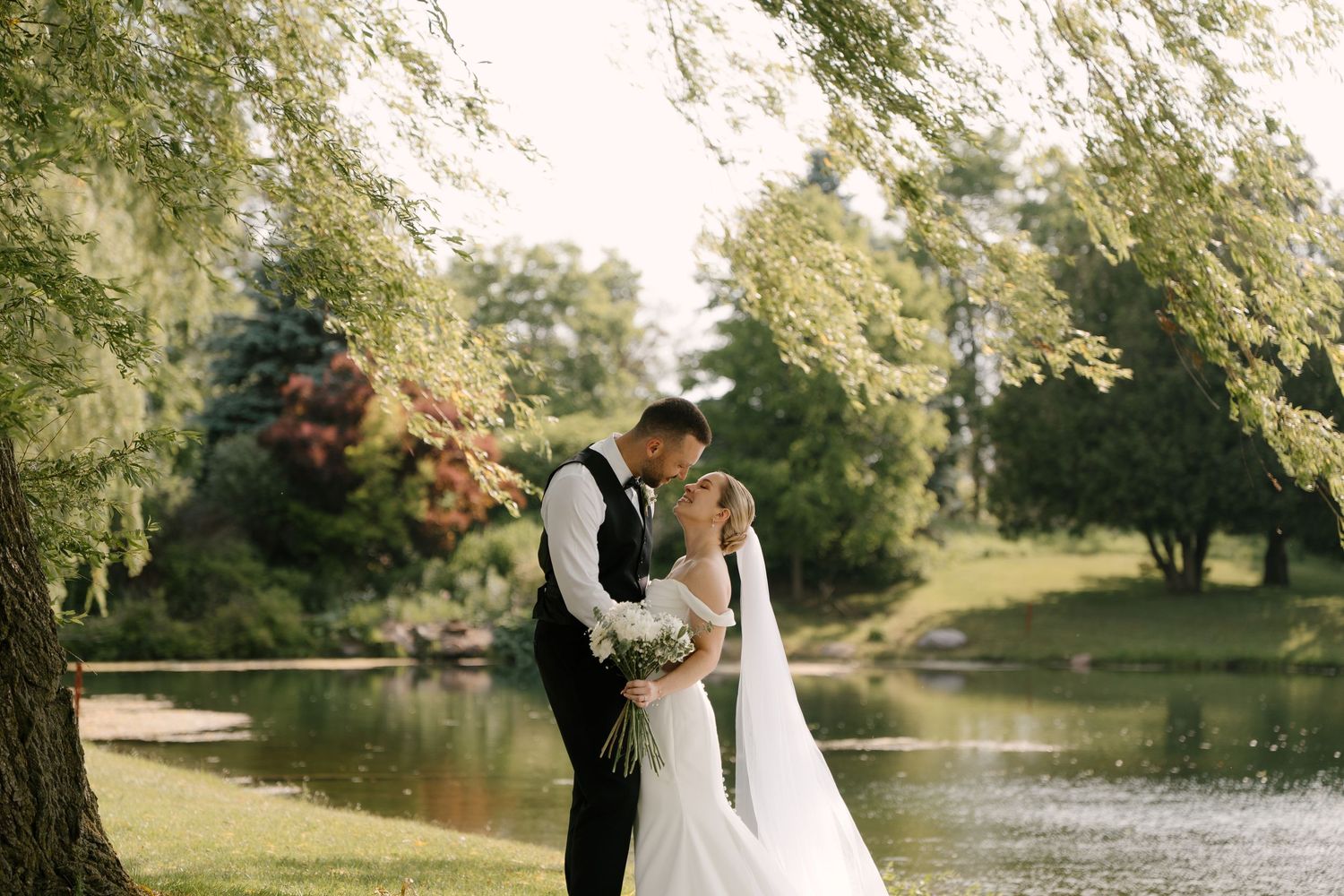 Wedding couple shares a romantic kiss near a tranquil lake surrounded by lush greenery.