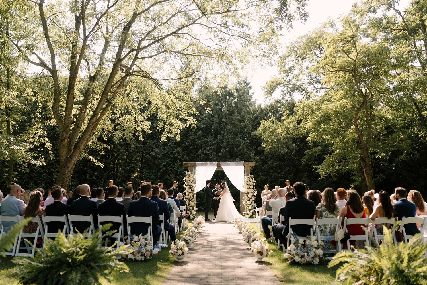 Outdoor wedding ceremony in a lush garden with guests seated on white chairs facing a floral arch and couple at the altar.