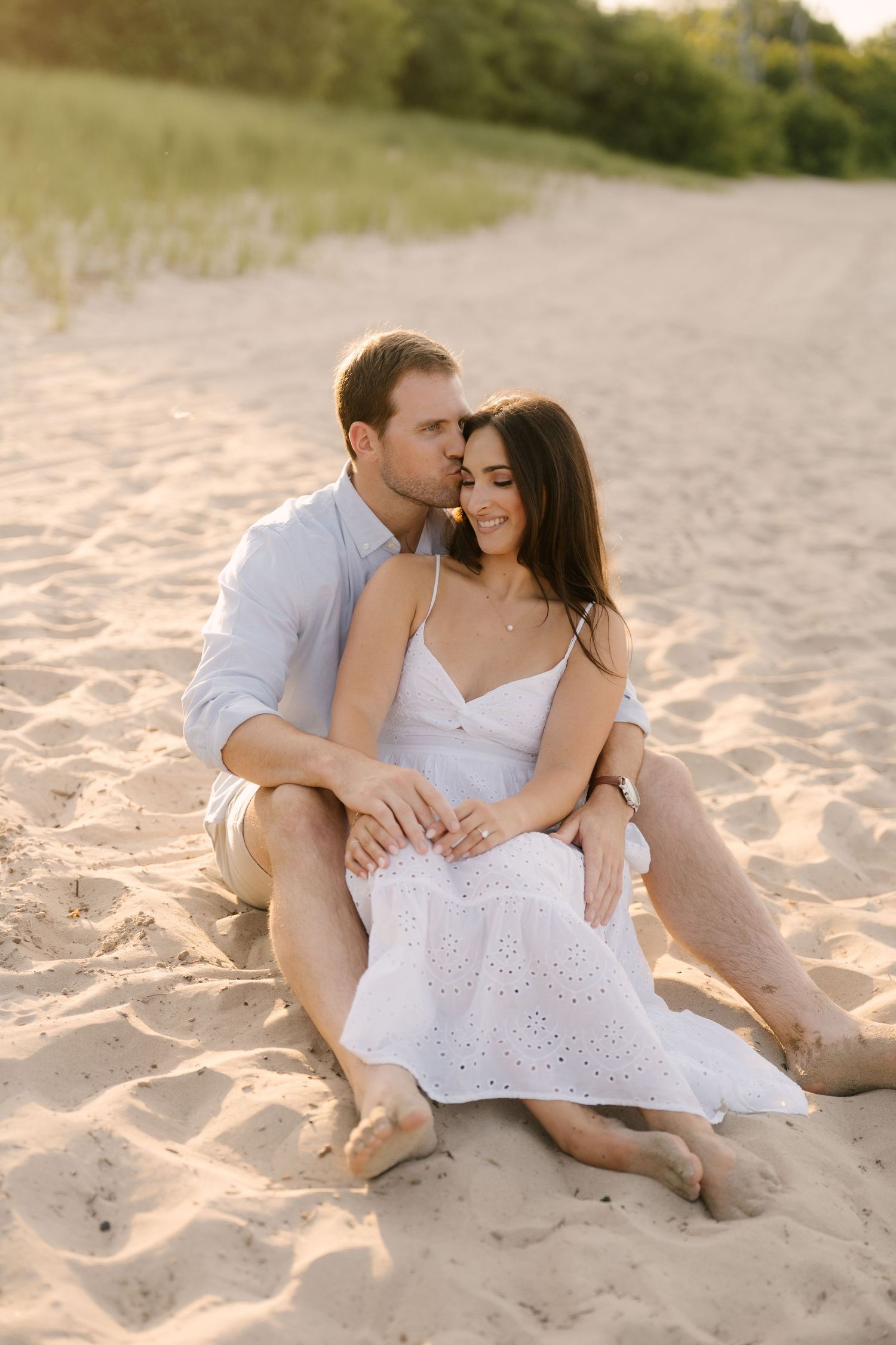 Romantic couple sitting together on sandy beach at sunset, man kissing womans temple.