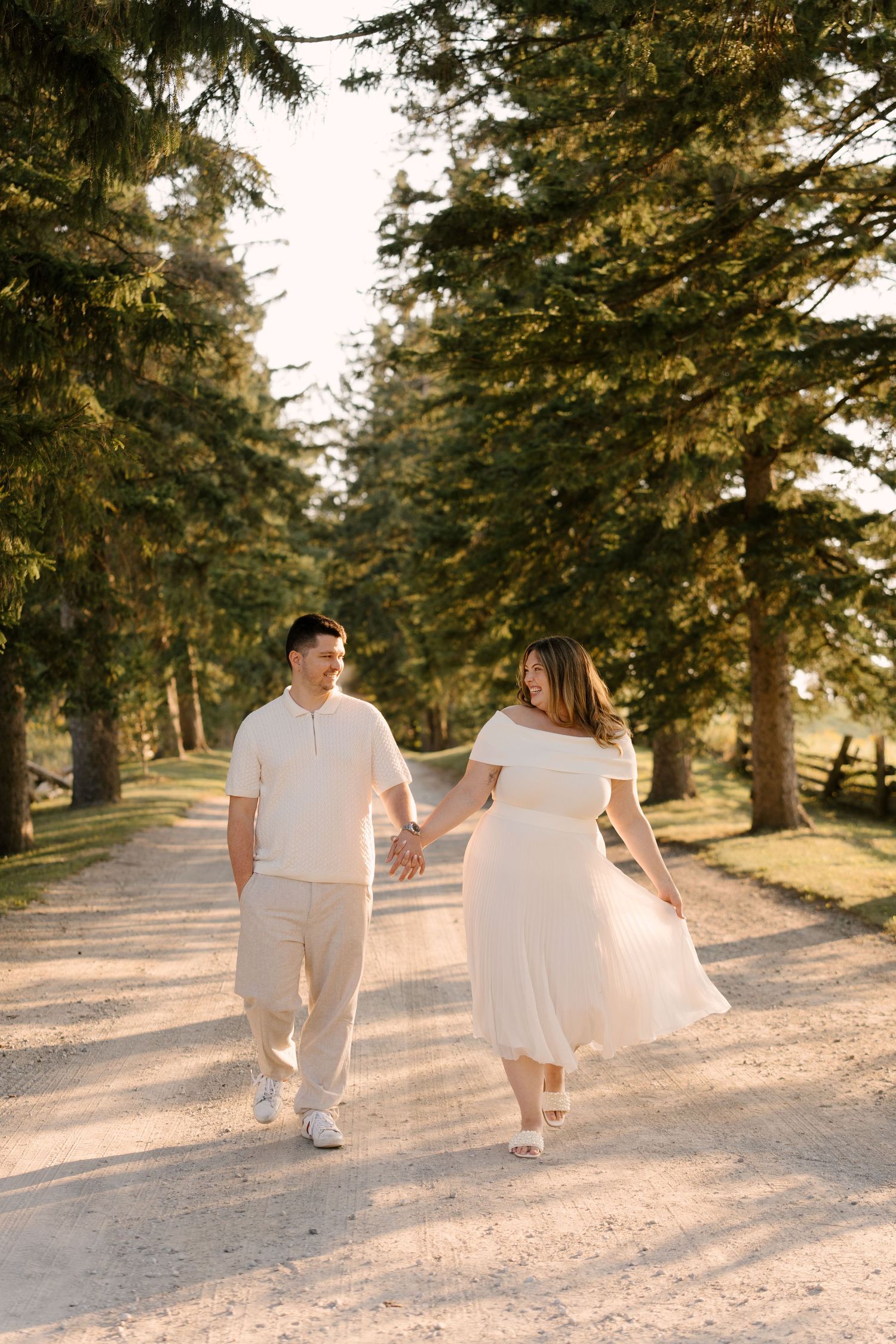 Couple in coordinating cream outfits holding hands and walking along a sun-dappled path lined with tall pine trees.
