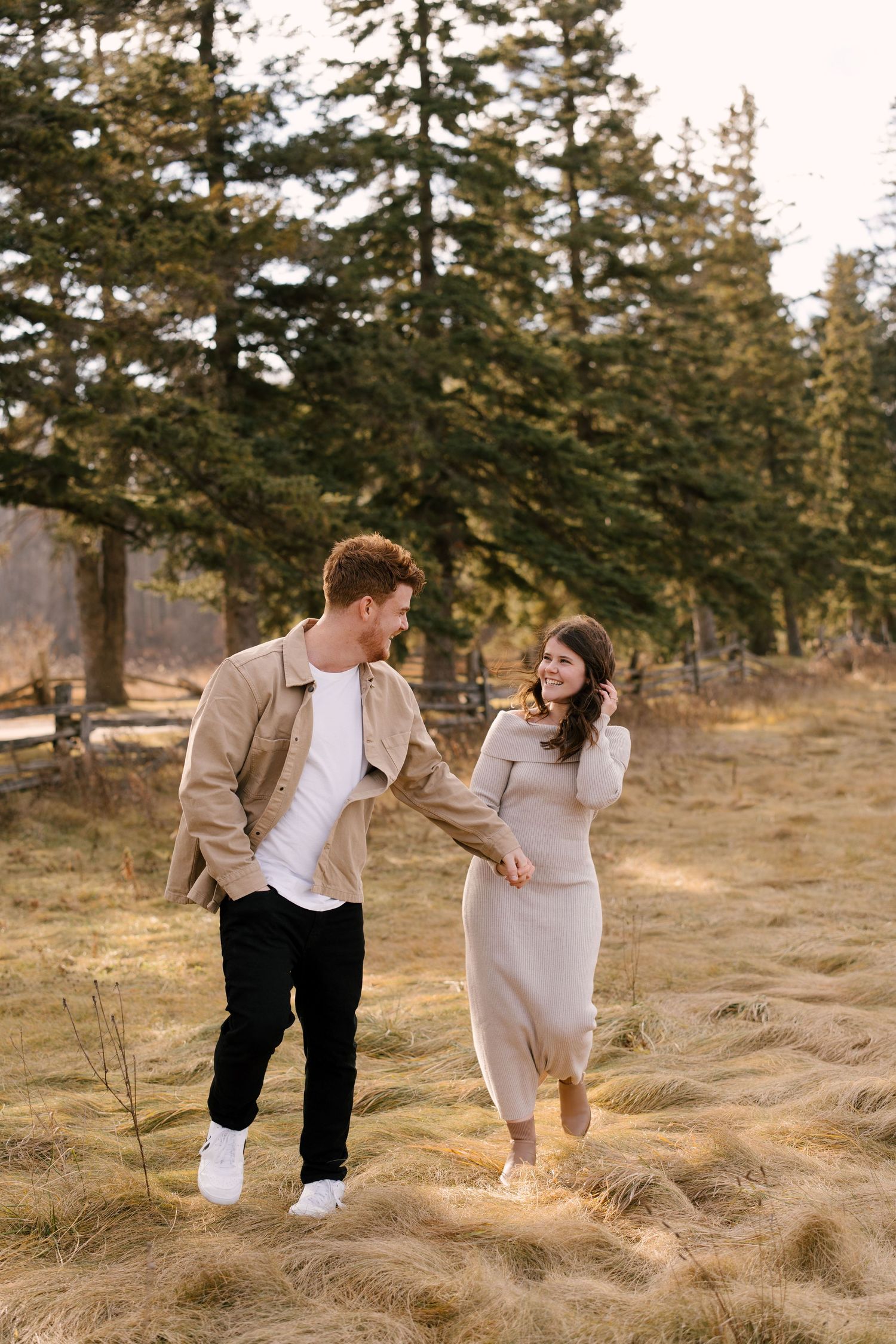 Couple holding hands and laughing while walking through a pine tree forest during golden hour.