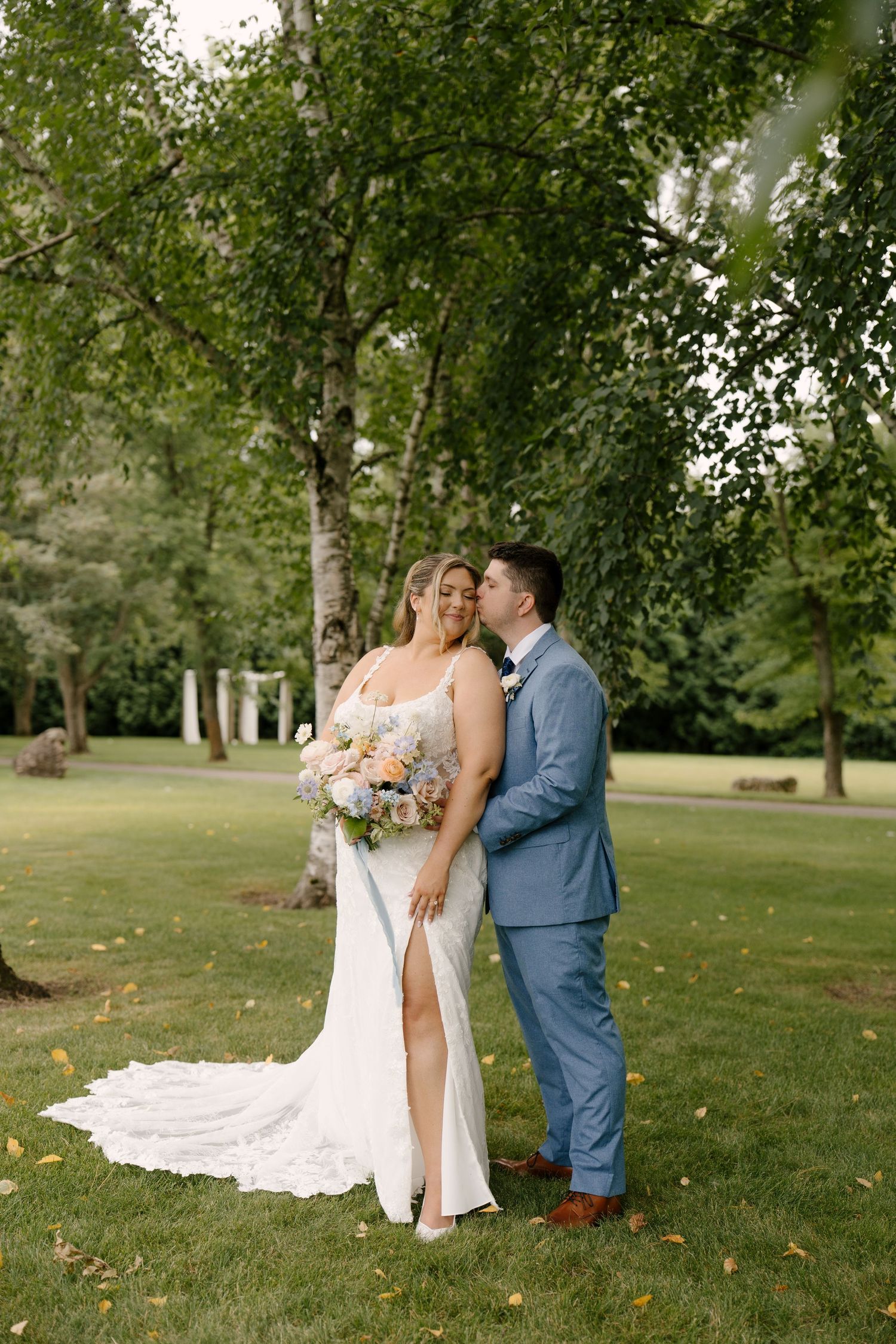 Groom in blue suit kissing bride's cheek as she smiles holding pastel bouquet under birch trees outdoors at Roseville Estates.