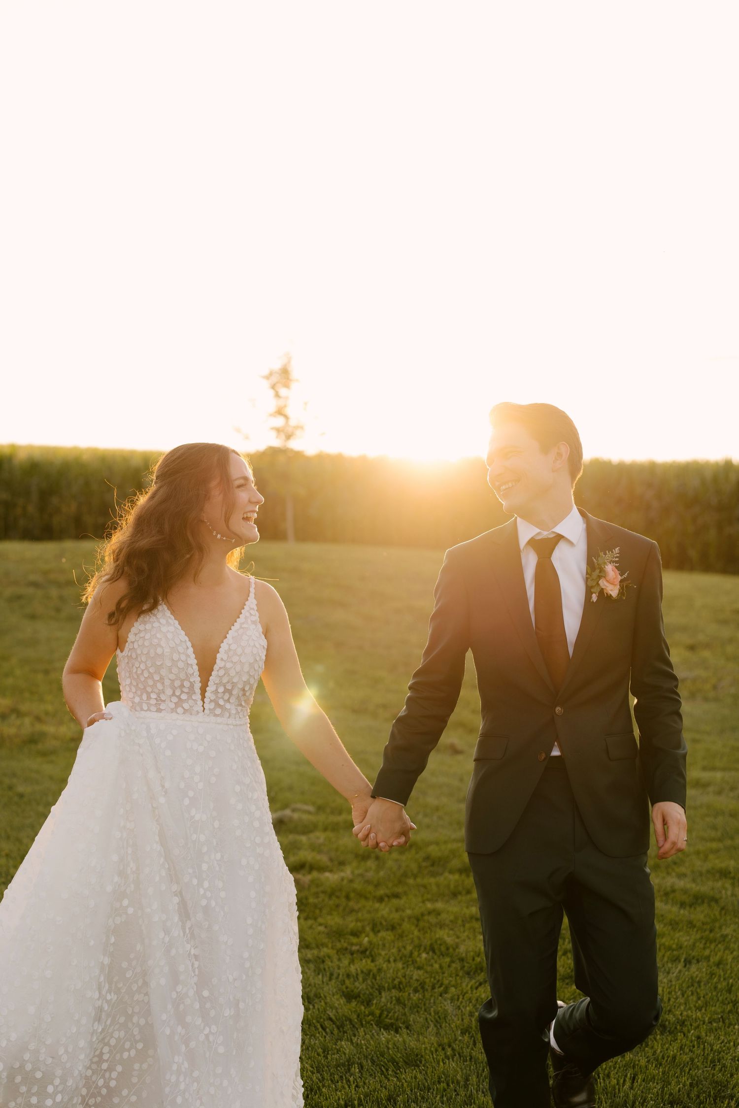 Bride and groom hold hands walking through golden sunset light in a lush green field at Wakefield Estates.