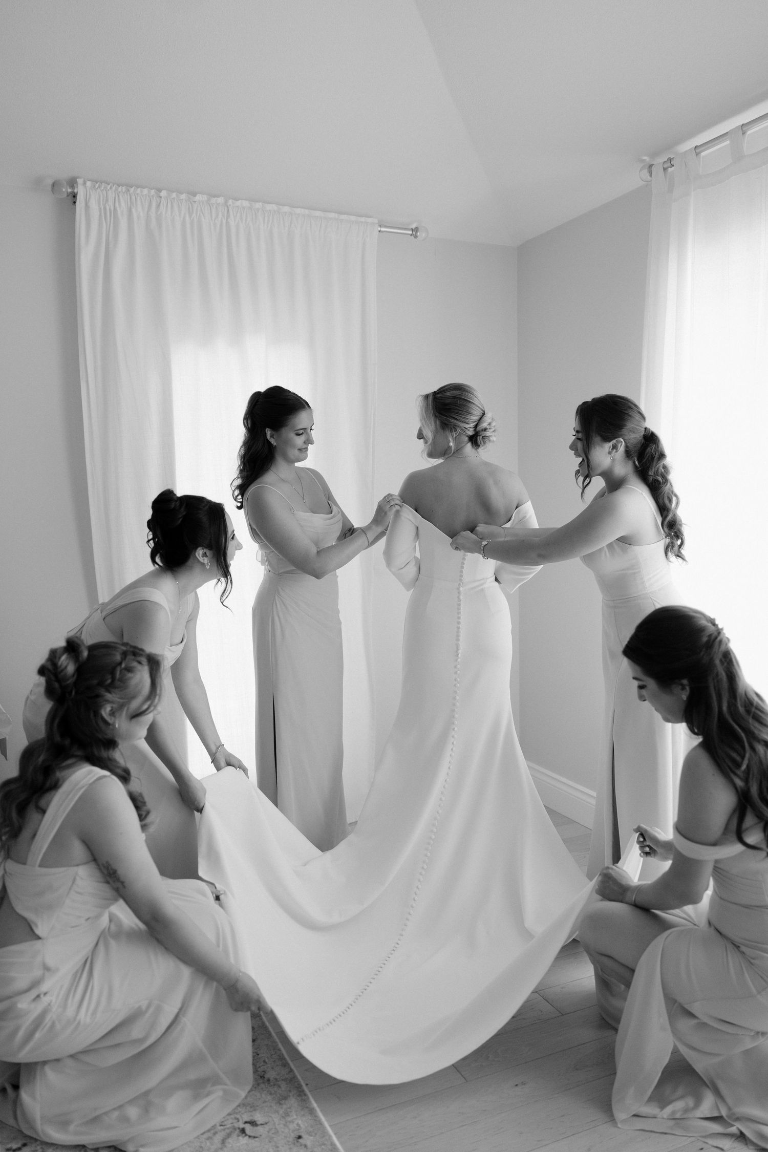 Black and white photo of bridesmaids helping bride into white wedding gown in bright windowed dressing room.