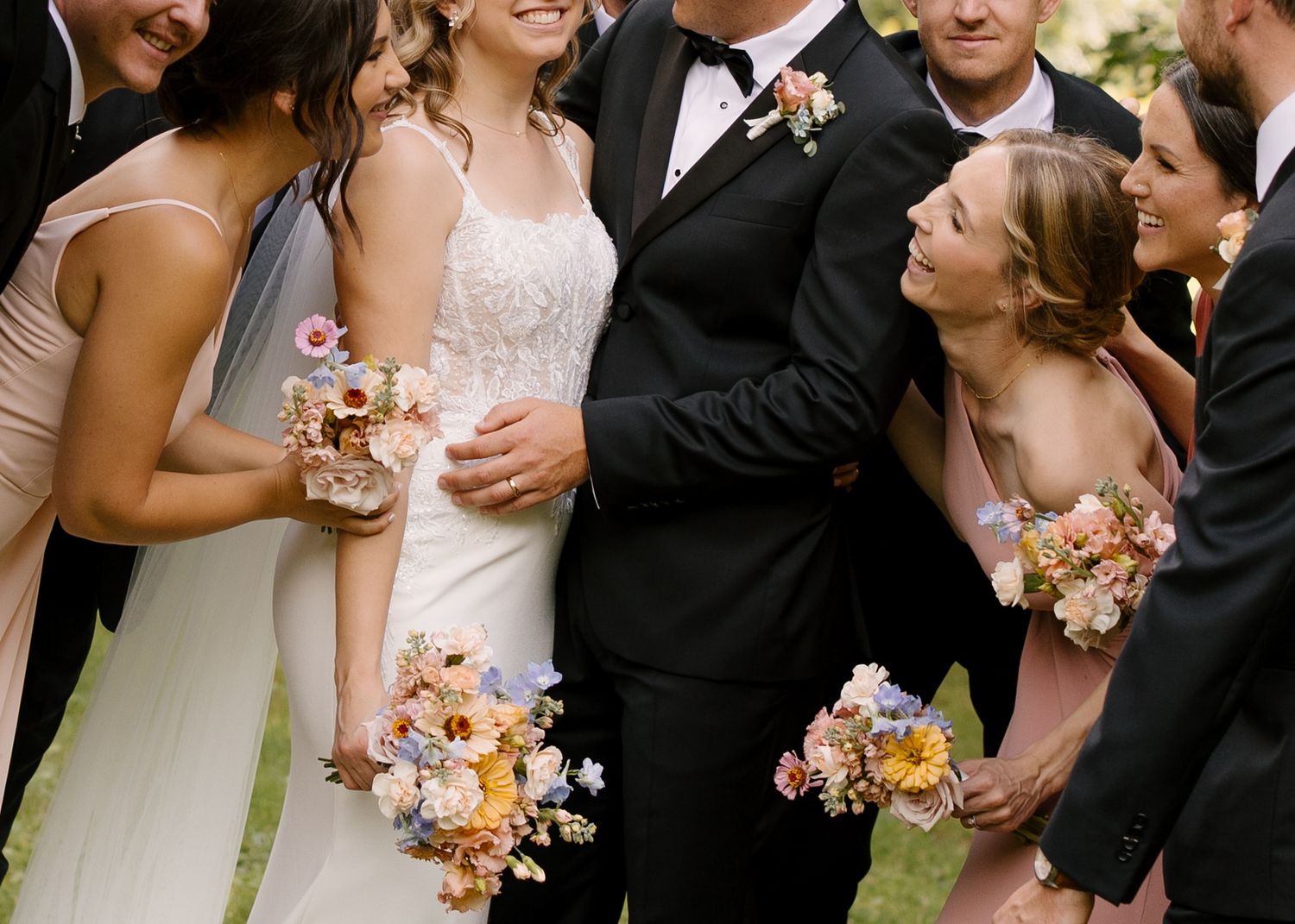 Bride and groom laughing joyfully with wedding party holding colorful bouquets of sunflowers and wildflowers.
