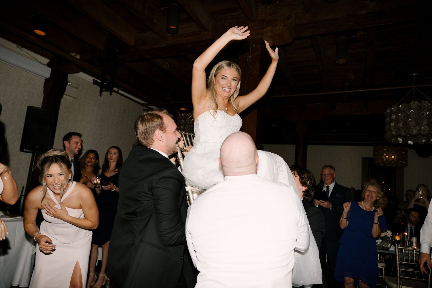 Joyful bride in white gown is lifted on a chair by guests during lively wedding reception celebration. At Ancaster Mill venue.