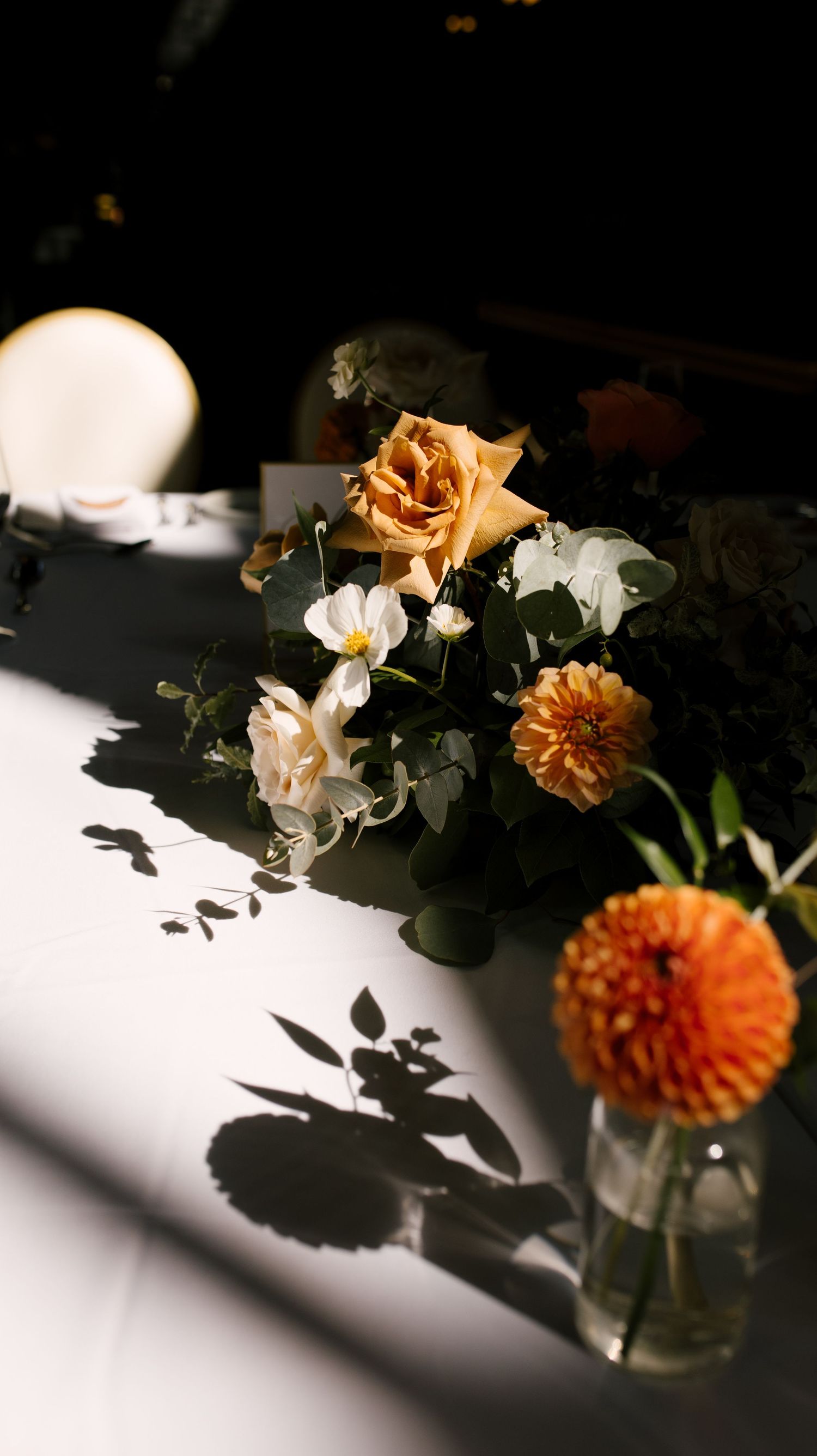Dramatic close-up of orange roses and white flowers casting shadows on a white tablecloth in moody lighting.