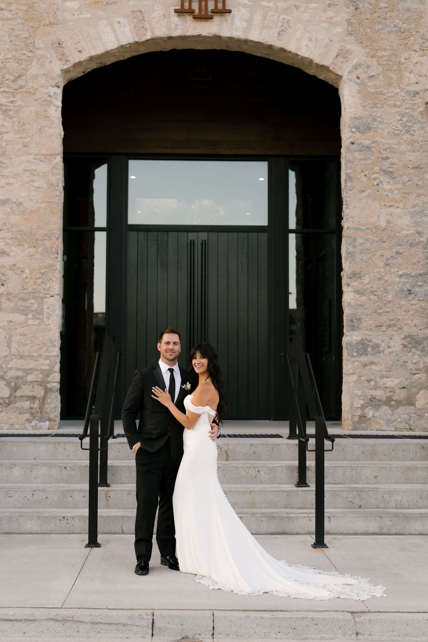 Bride and groom posing elegantly before large black arched doors of Tapestry Hall stone building.