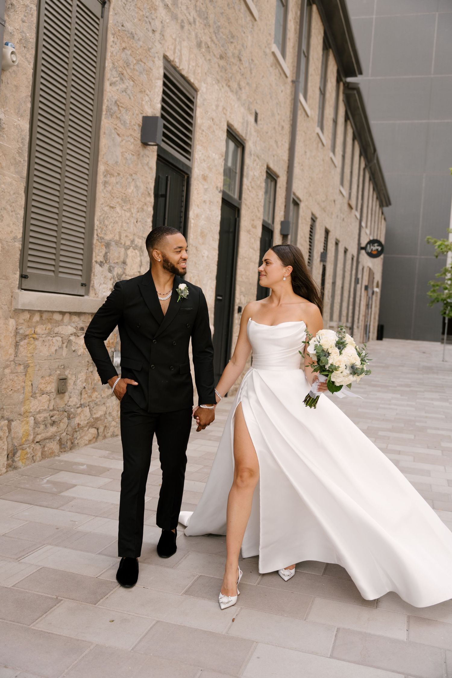 Bride and groom walk hand in hand along a stone building pathway on their wedding day in Cambridge Ontario.