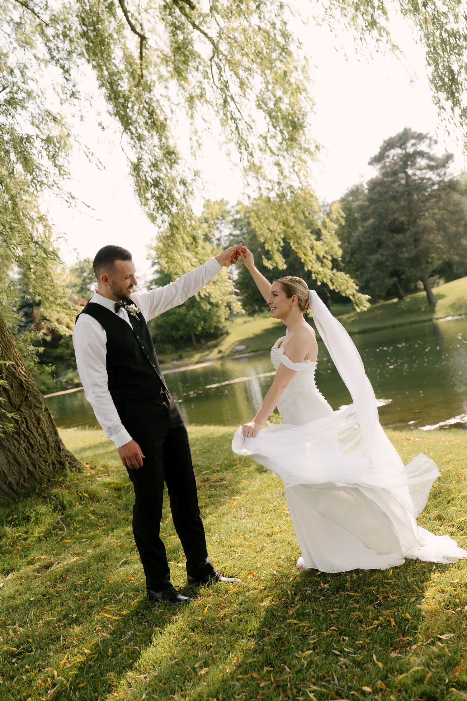 Groom spins bride in her elegant white wedding dress beside a sunlit lake and willow tree in Guelph.