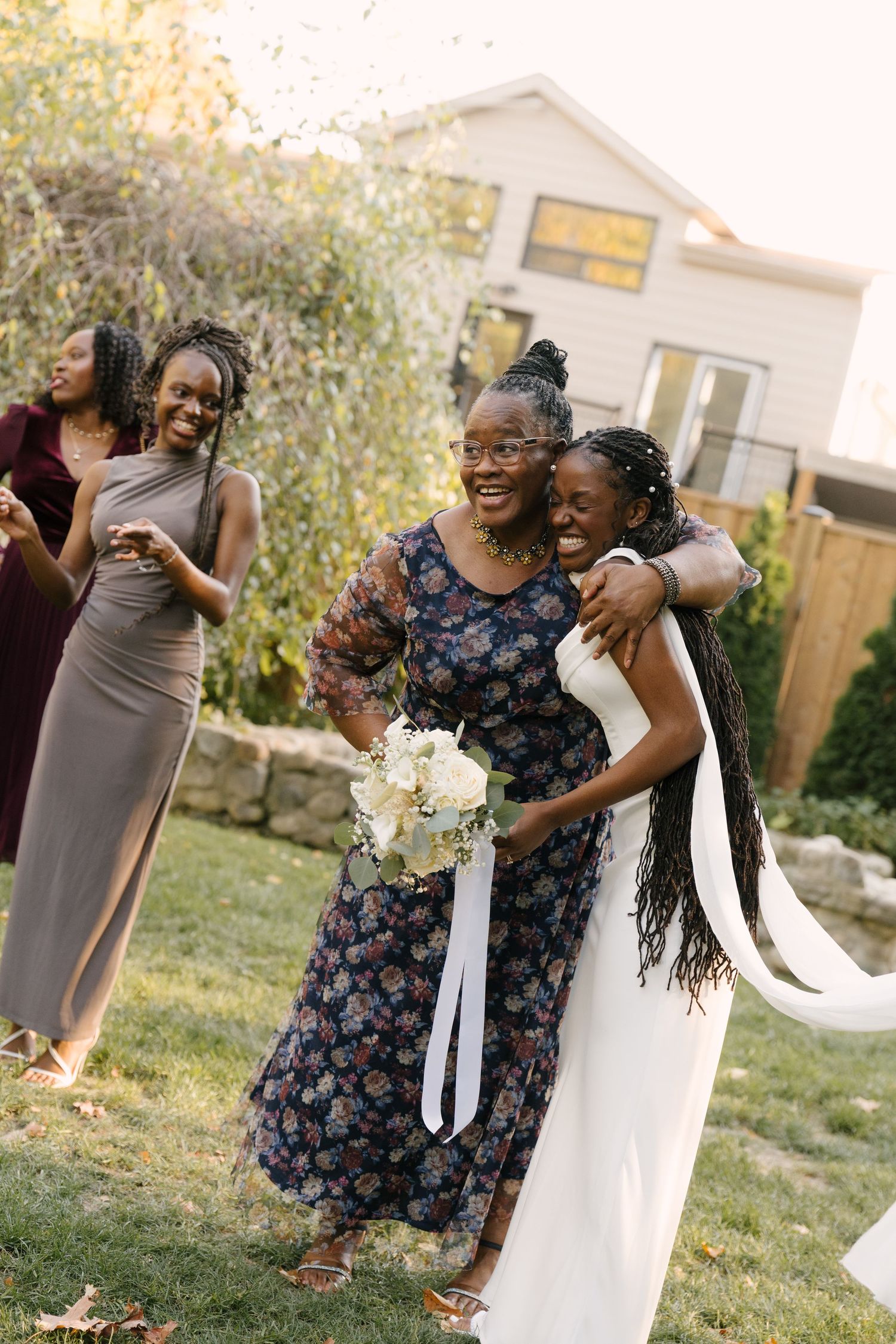 A bride embraces an older woman in a floral dress at an outdoor wedding celebration with guests in the background.