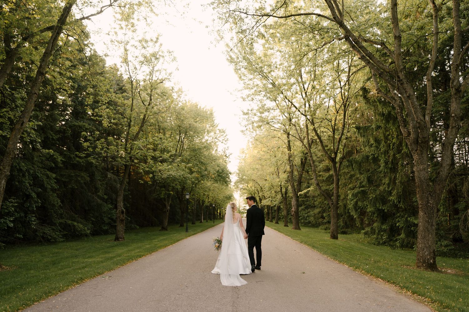 Bride and groom holding hands on a tree-lined path surrounded by lush green foliage on their wedding day.