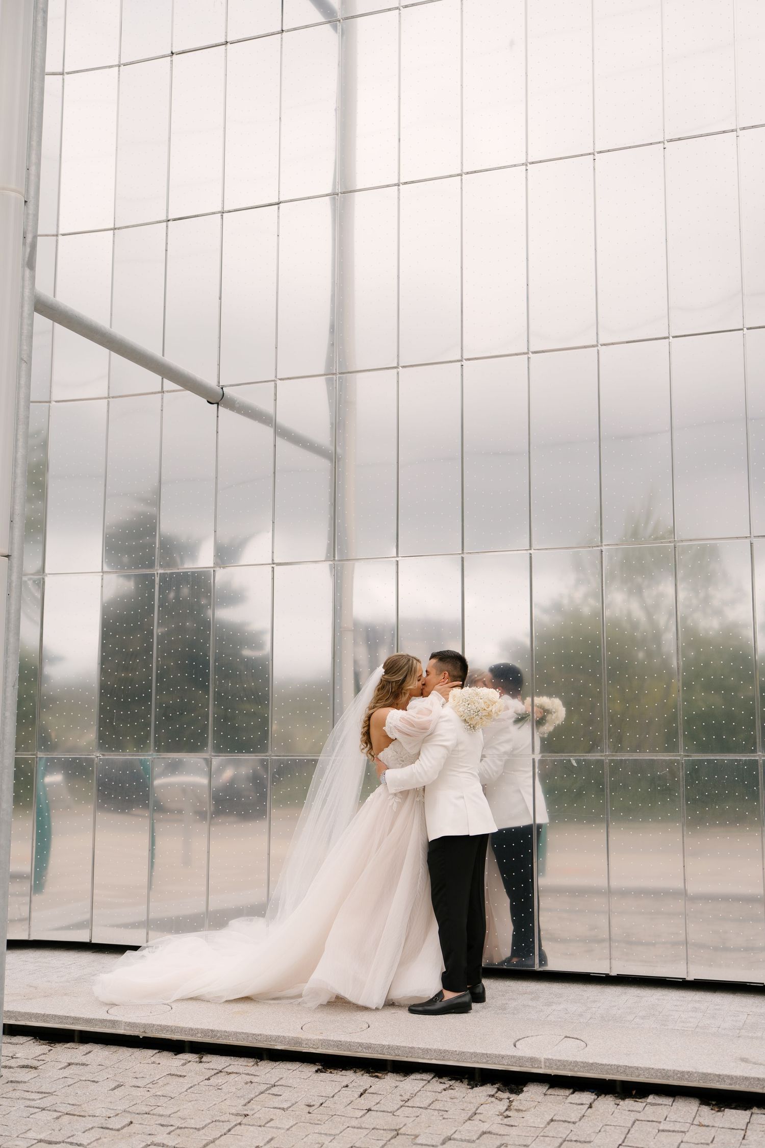 Bride and groom share a romantic kiss in front of a large glass building facade.
