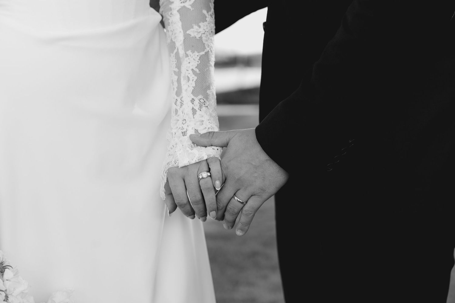 Close-up of bride and groom holding hands showing wedding rings, lace wedding dress detail.