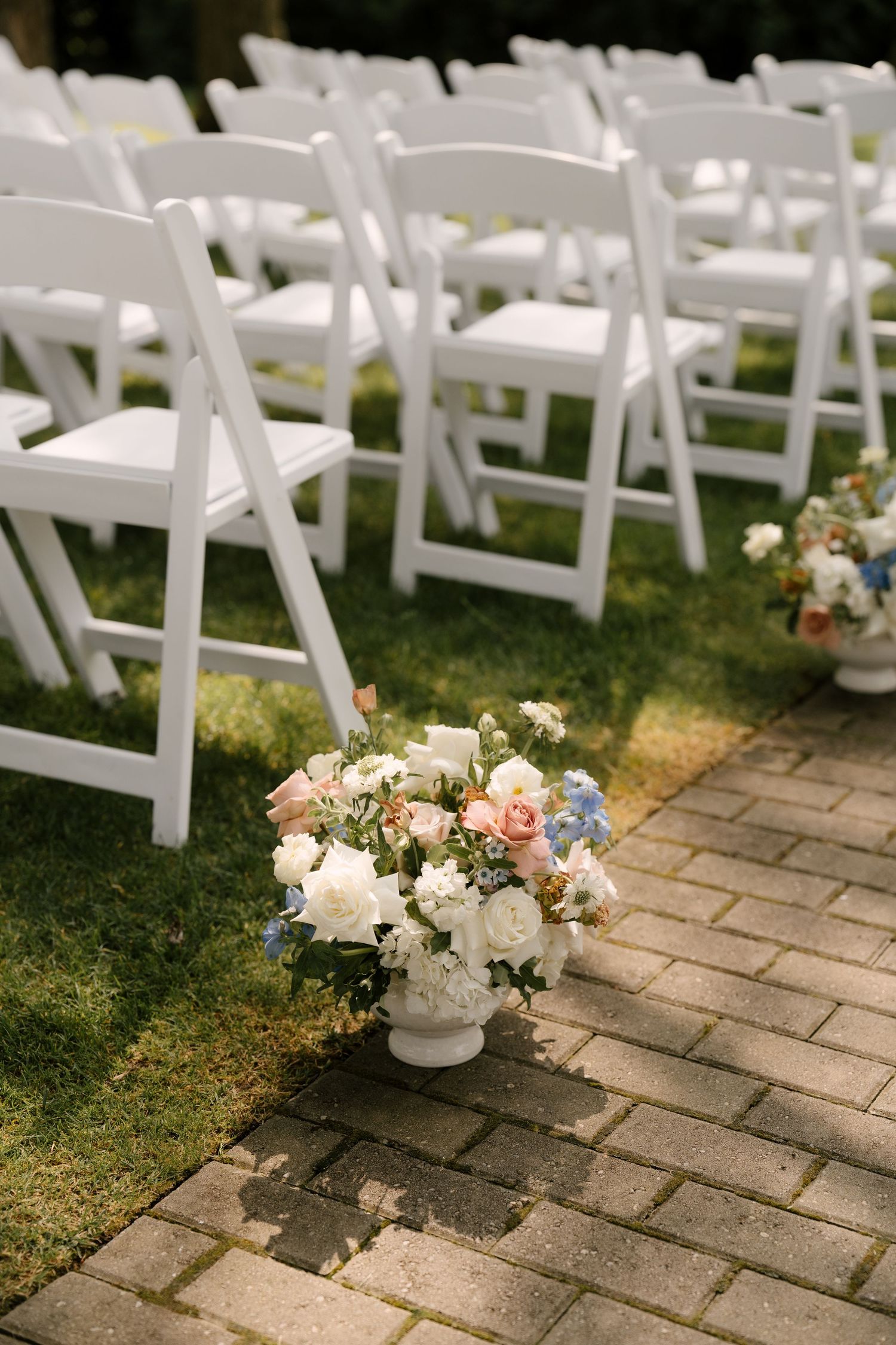 White urn filled with pink, blue, and white roses decorating the brick aisle at an outdoor wedding ceremony.