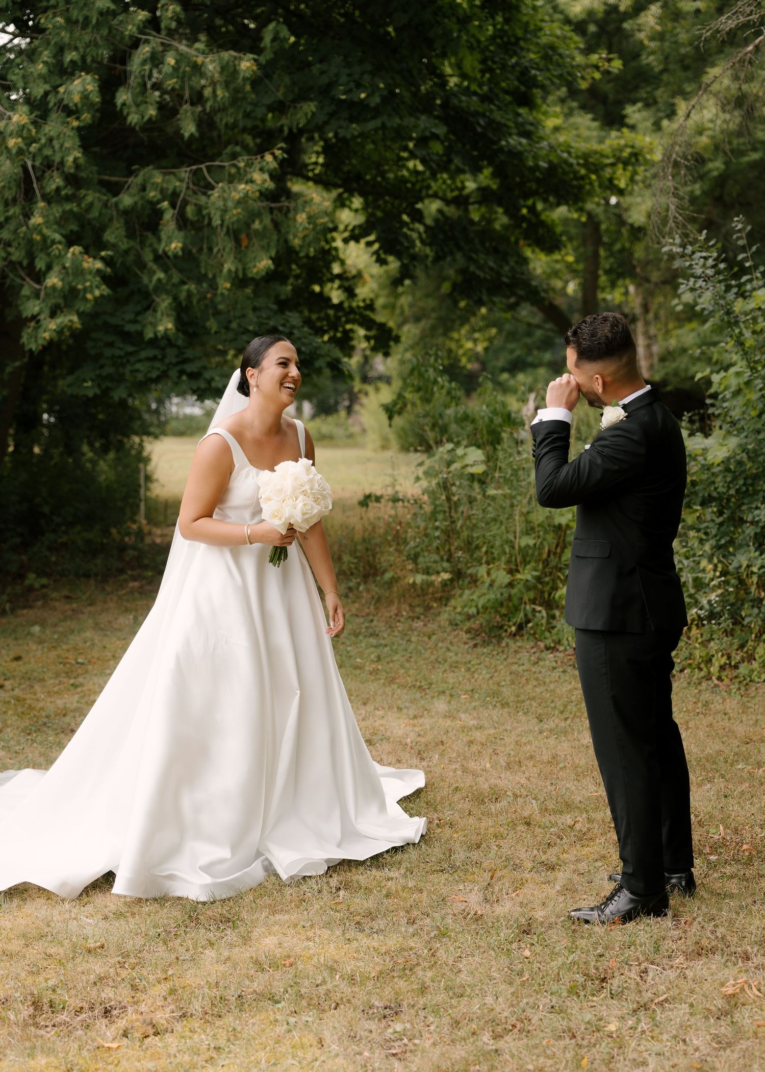 Emotional groom wipes tears seeing bride in white gown for first time during outdoor wedding first look.