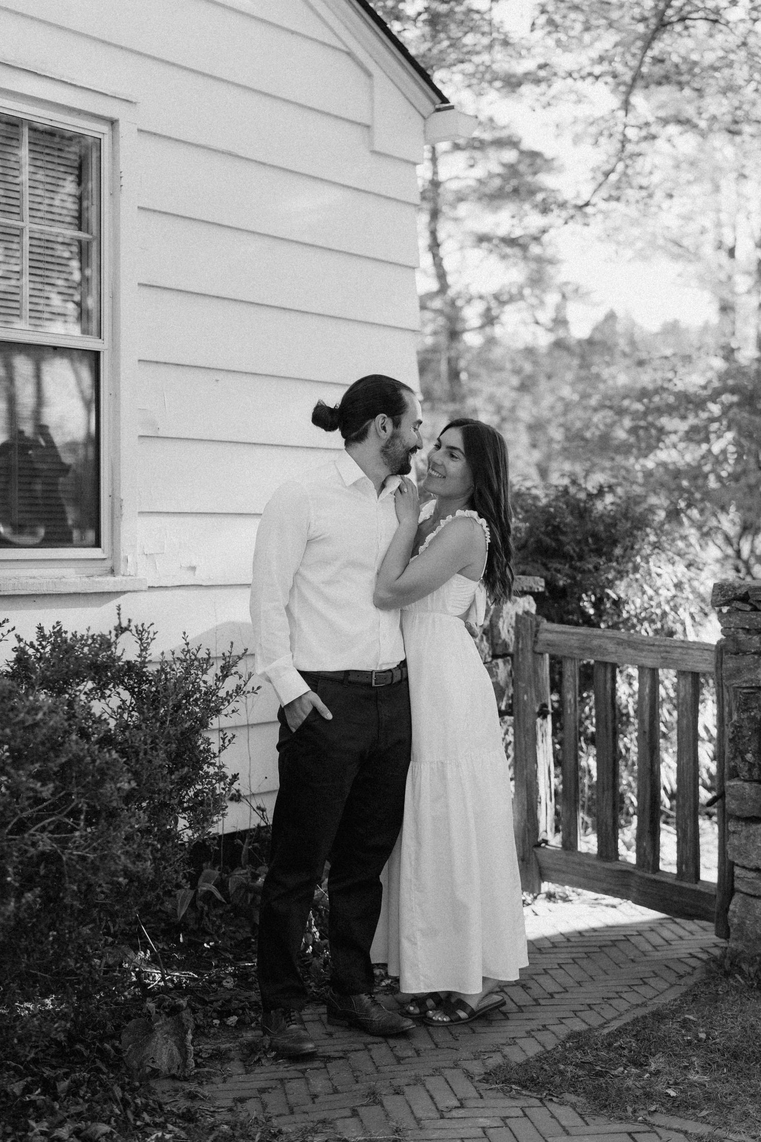 Black and white photo of a couple embracing romantically outside a white cottage with wooden fence.