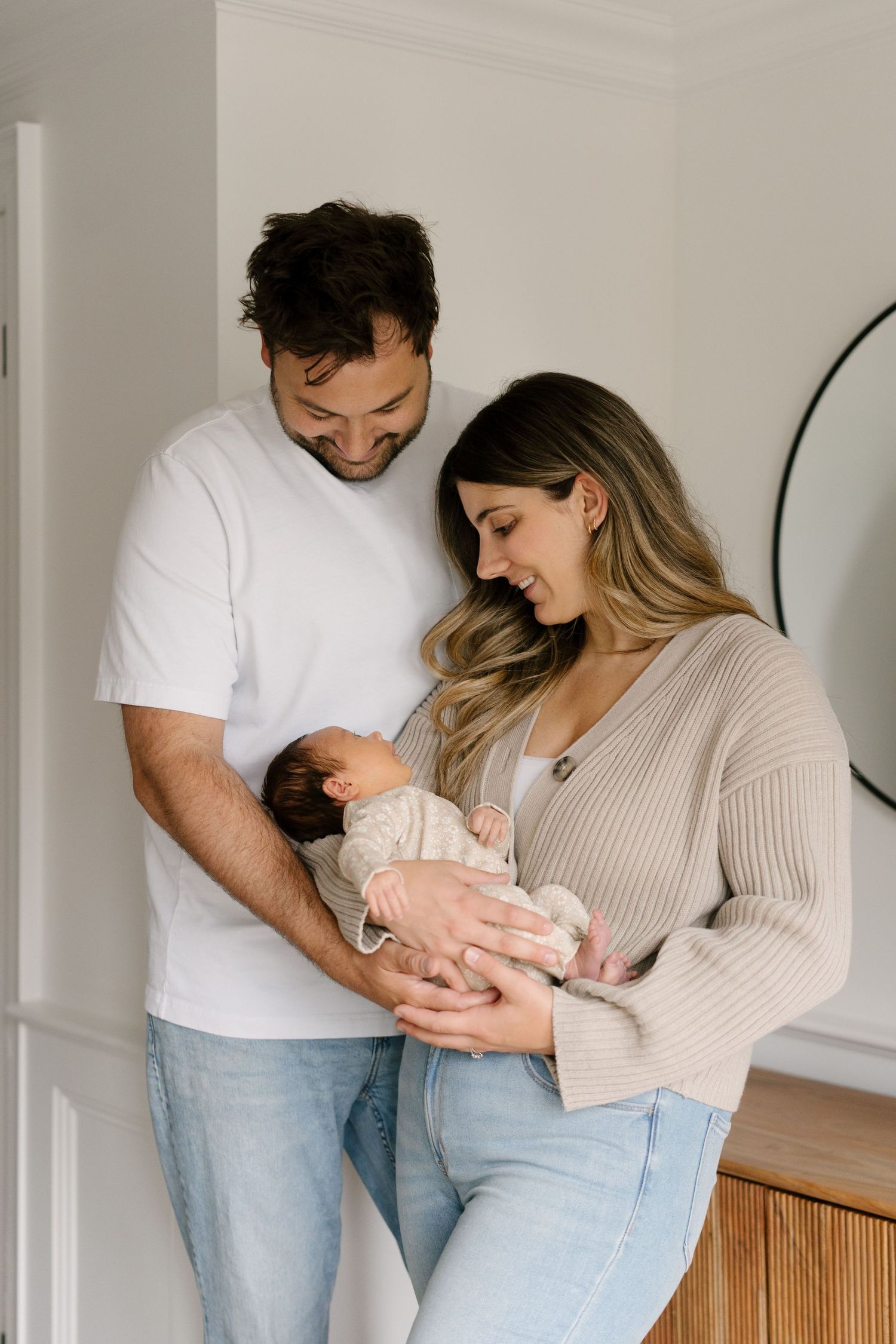Happy couple in casual attire smiling at their newborn baby in a bright, modern home setting.
