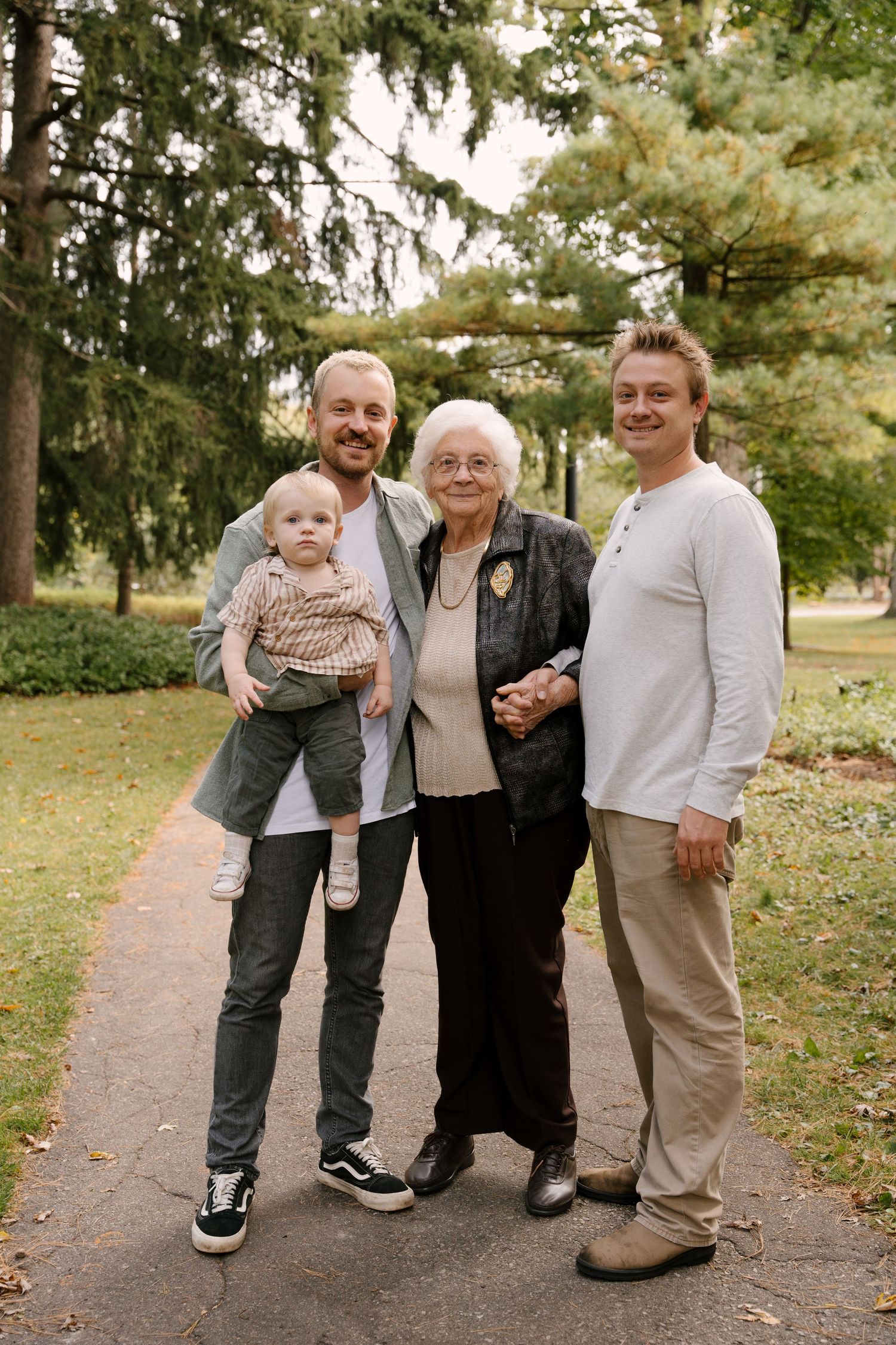Multi-generational family portrait with two men, elderly woman, and baby posing on park path.